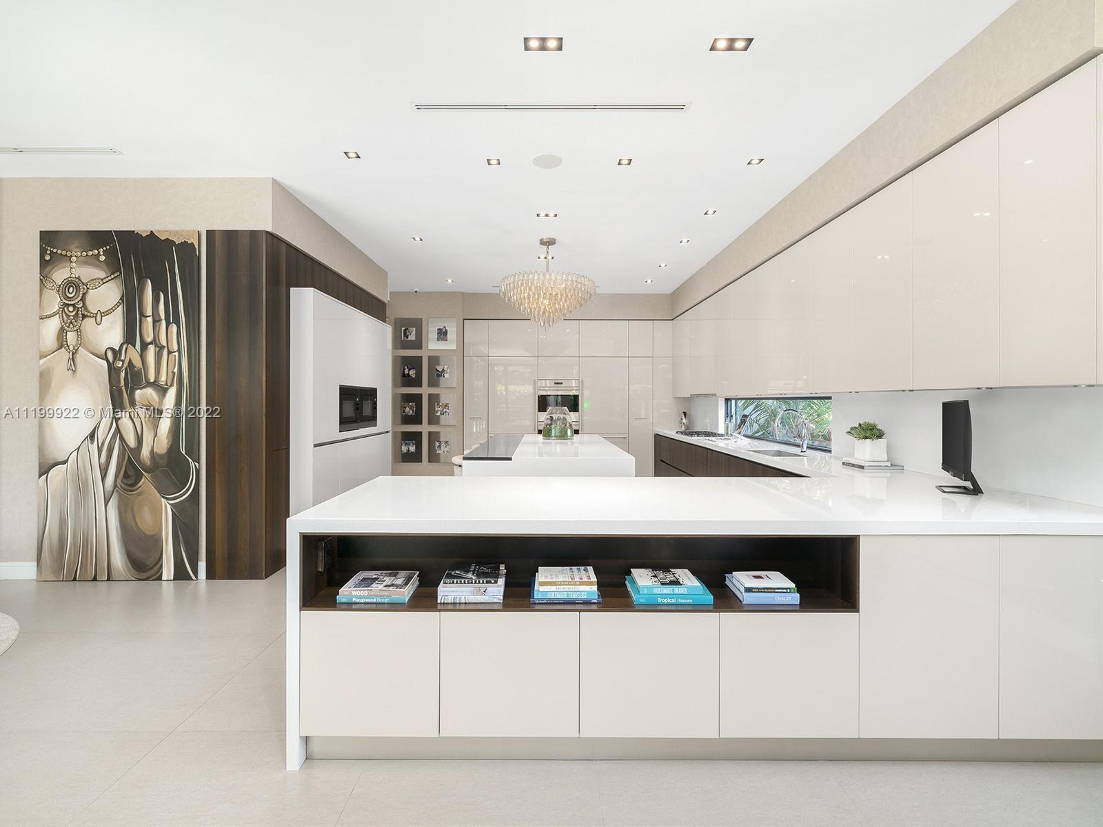 Modern white kitchen with a large island, glossy cabinets, dark wood accents, and a crystal chandelier.