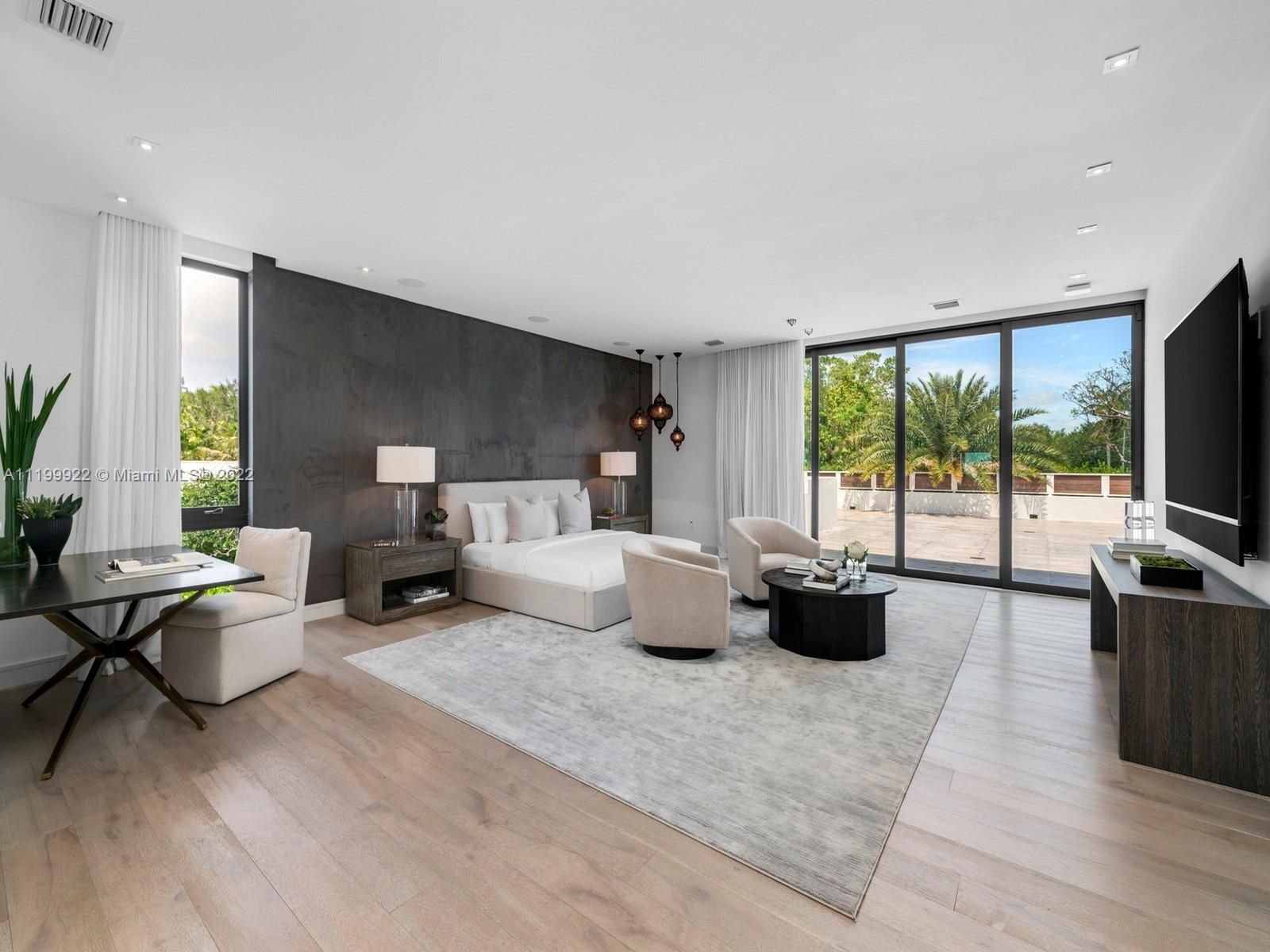 Modern bedroom with a dark accent wall, white bed, sitting area, and glass doors to a palm-lined terrace.