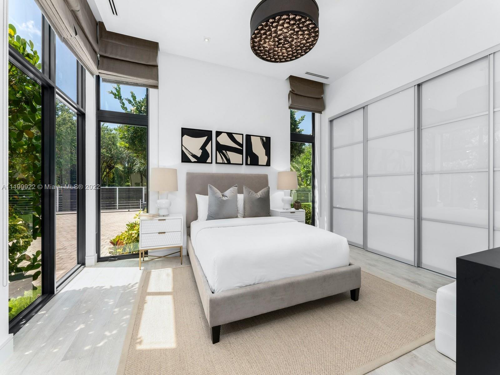 Bright modern bedroom with a gray upholstered bed, abstract art, floor-to-ceiling windows, and a large sliding-door closet.