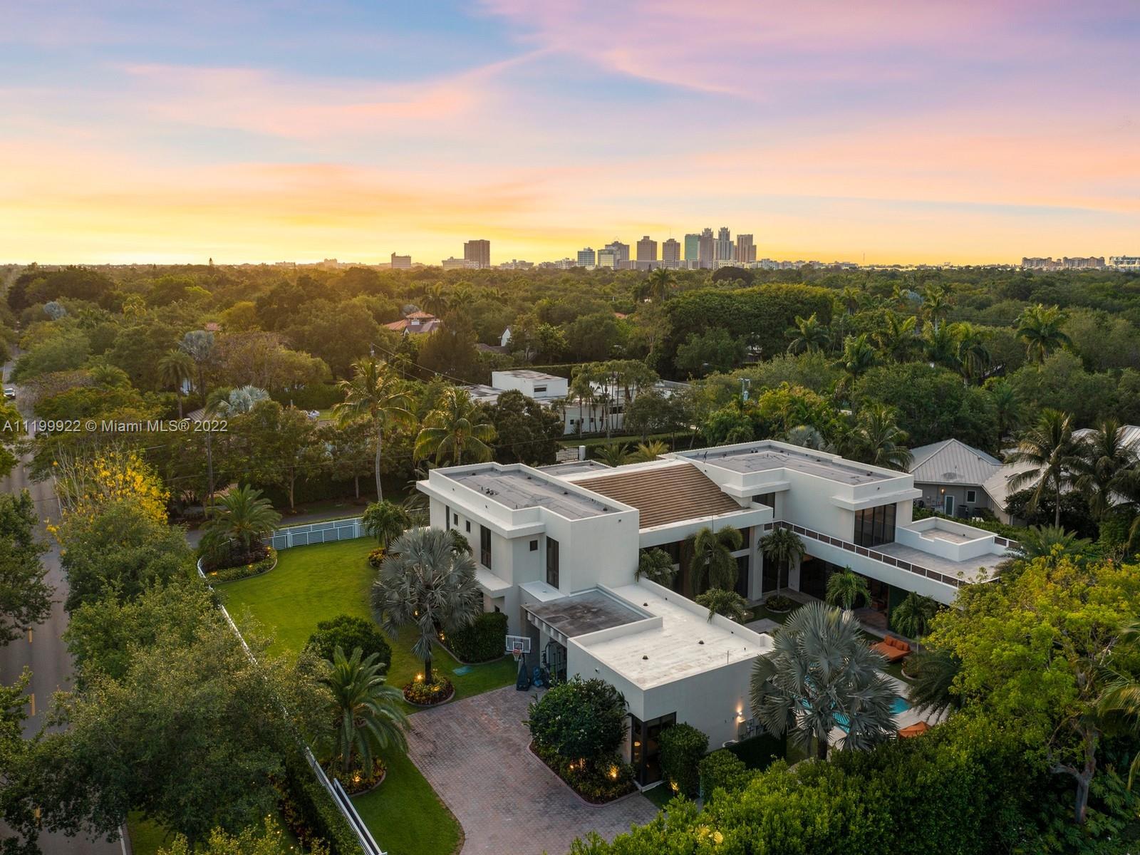 Aerial view of a modern white home and landscaped grounds, with a distant city skyline at sunset.