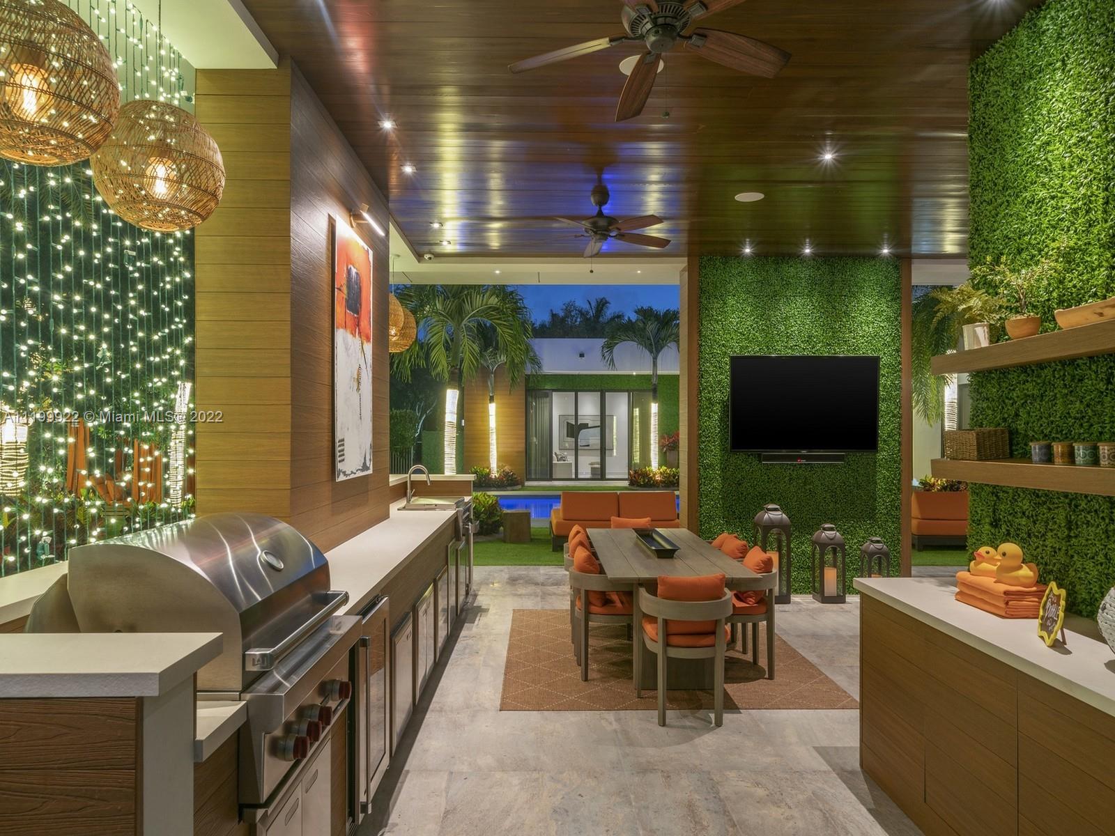 Covered outdoor patio with a built-in grill, dining set, hedge-wall television, and pool beyond.