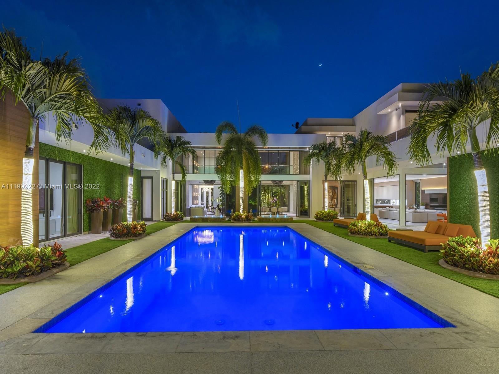 Illuminated pool in front of a modern home, framed by palms and outdoor seating at night.