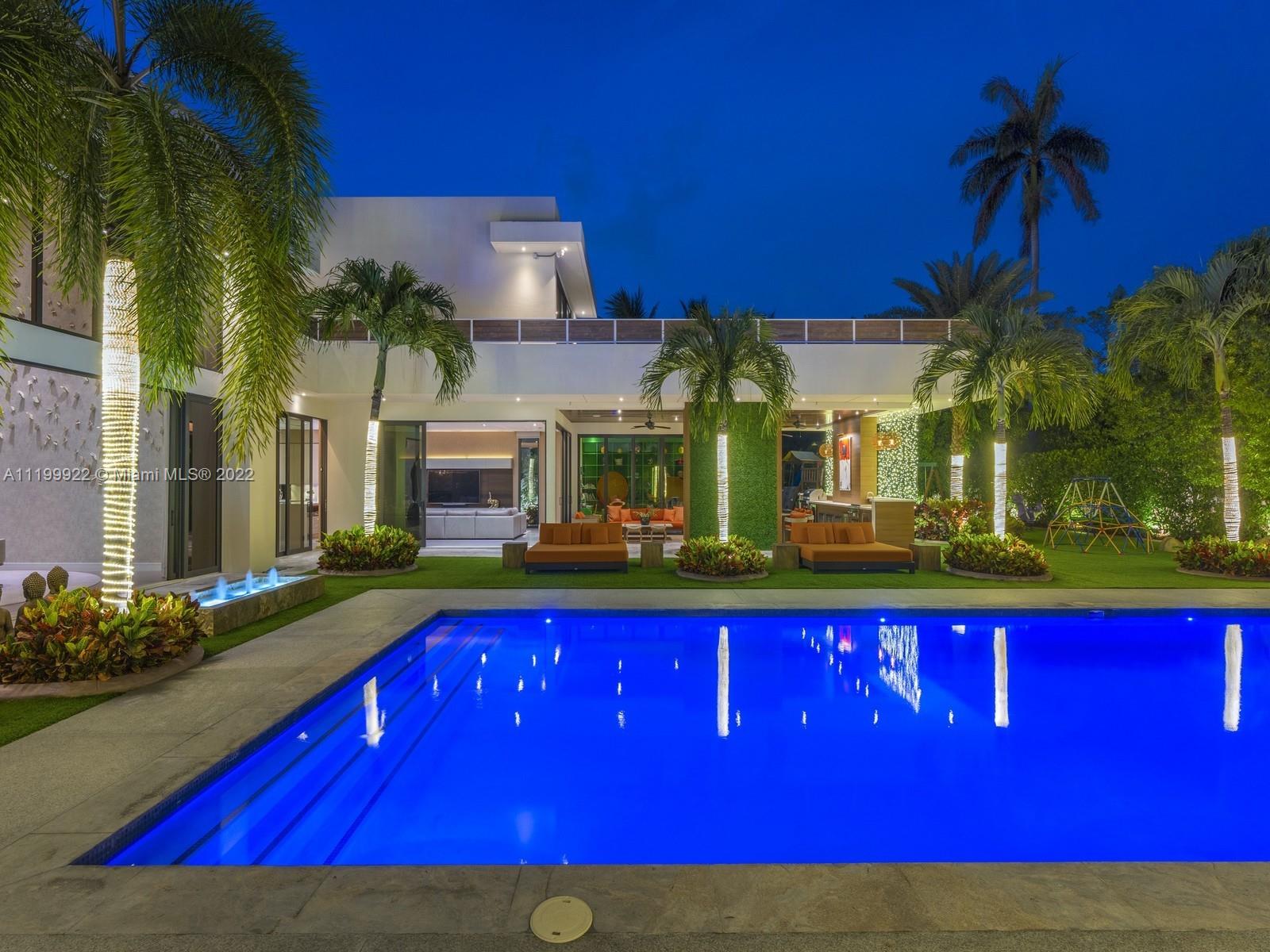 Modern home beside a brightly lit pool, with tropical palms and outdoor lounge seating.
