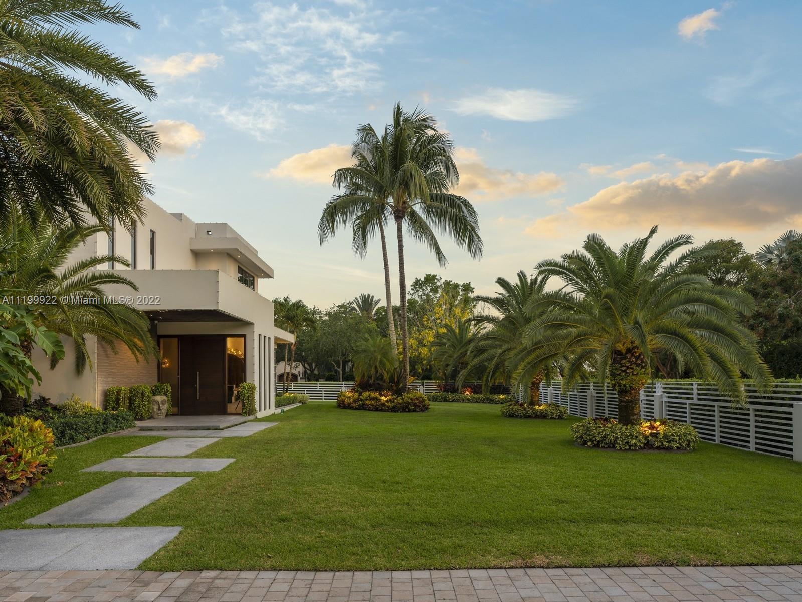 Modern white home with a broad lawn, tropical palms, landscaped beds, and a white fence.