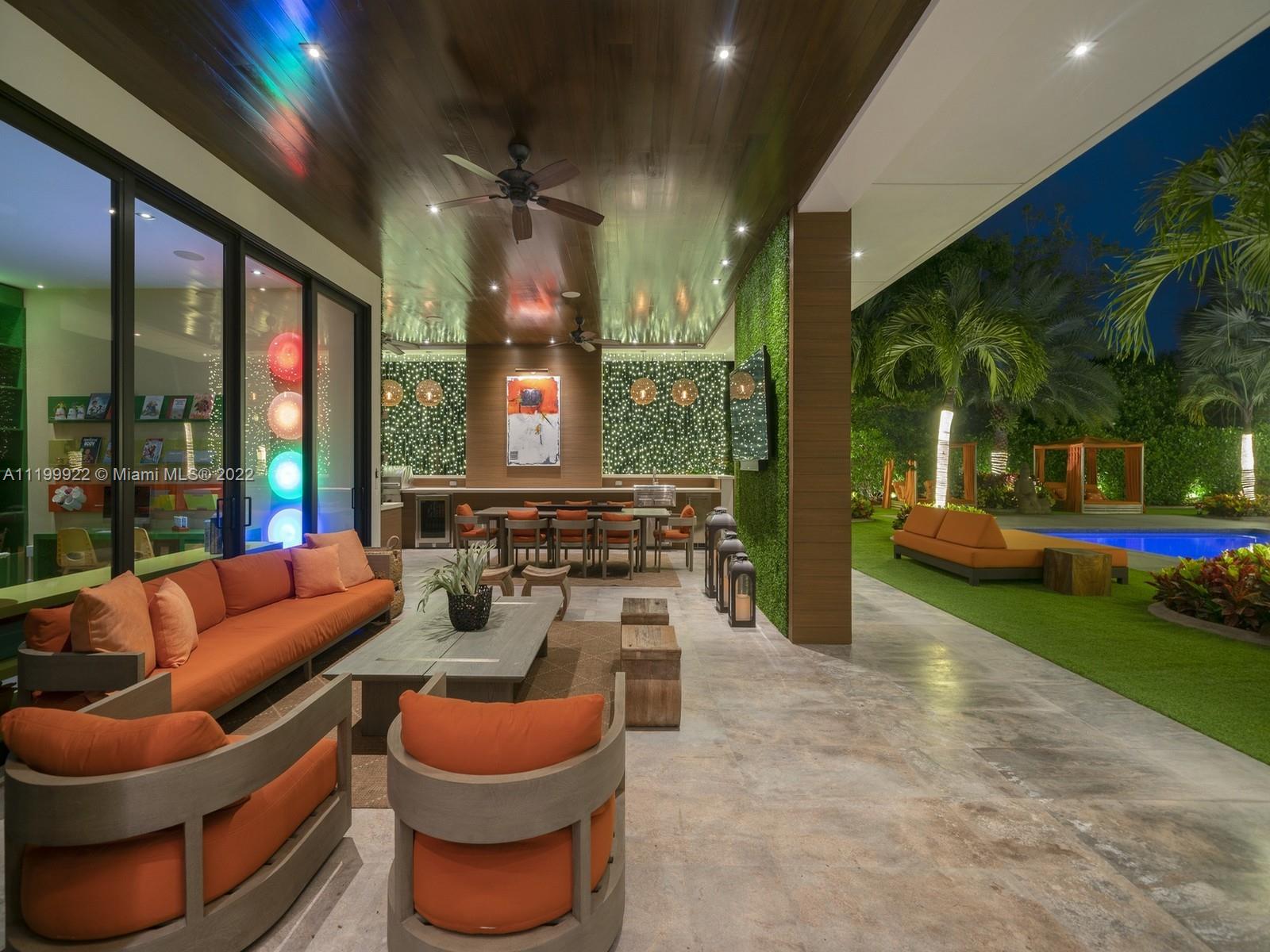 Covered patio with orange seating and dining area beside a lit pool and tropical landscaping.