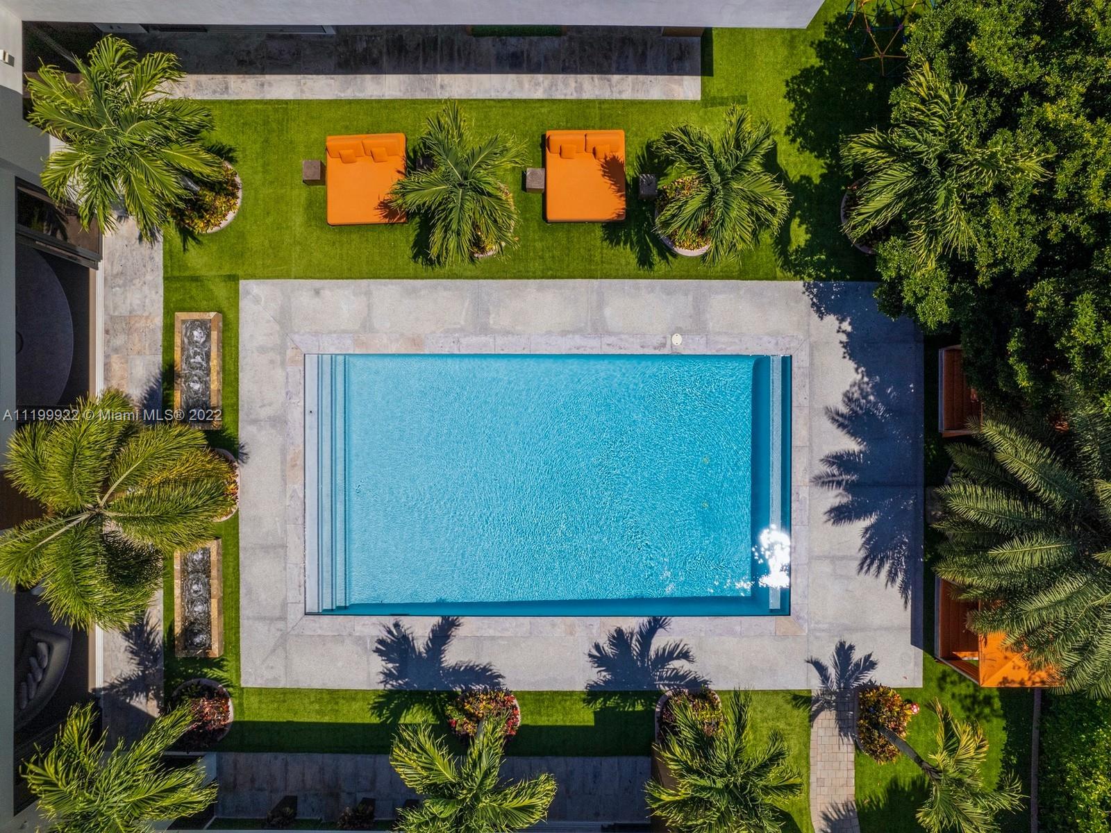 Aerial view of a backyard pool with pale stone decking, palm trees, and orange loungers.