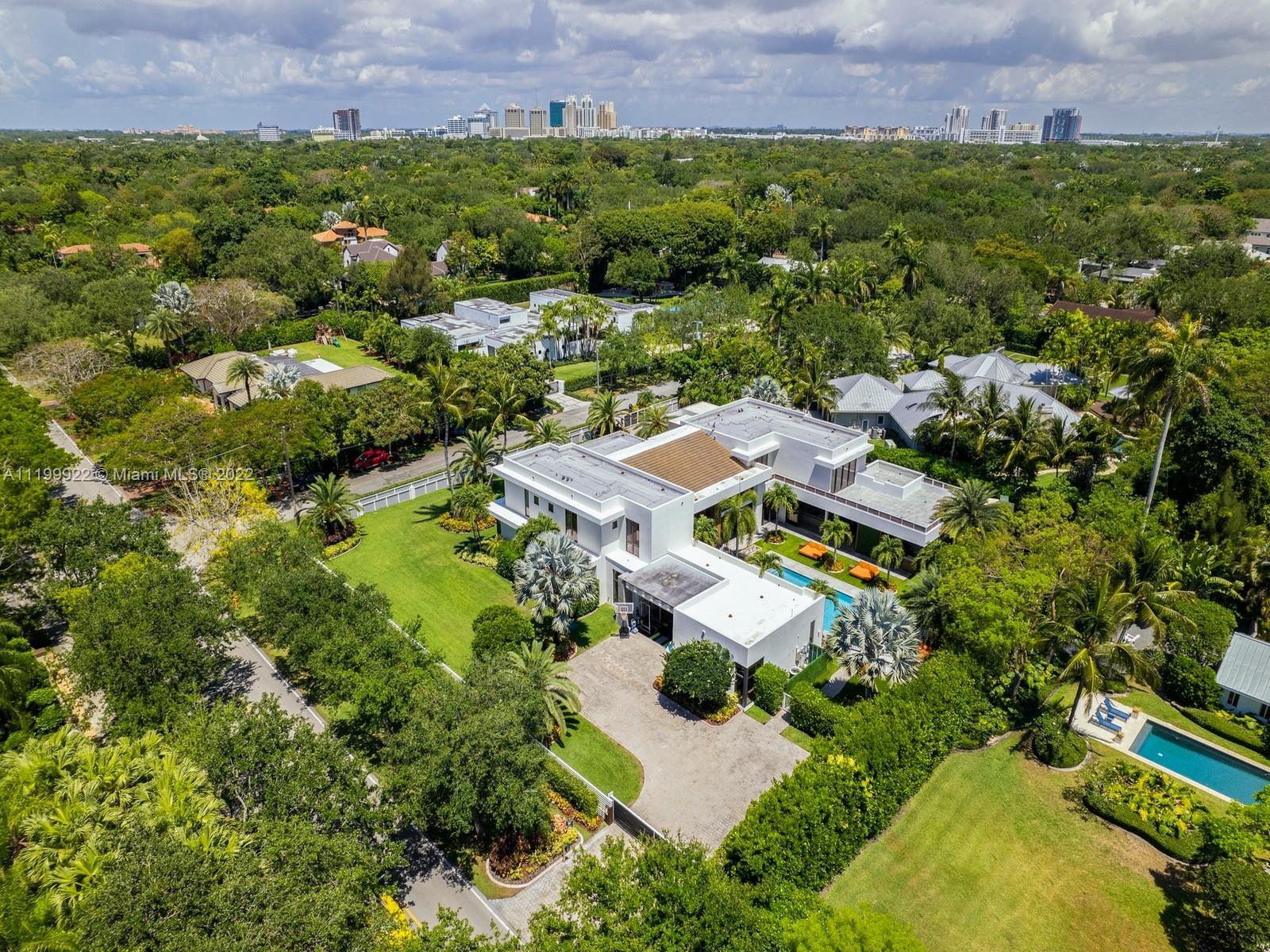 Aerial view of a modern white home with a pool, tropical landscaping, and distant skyline.