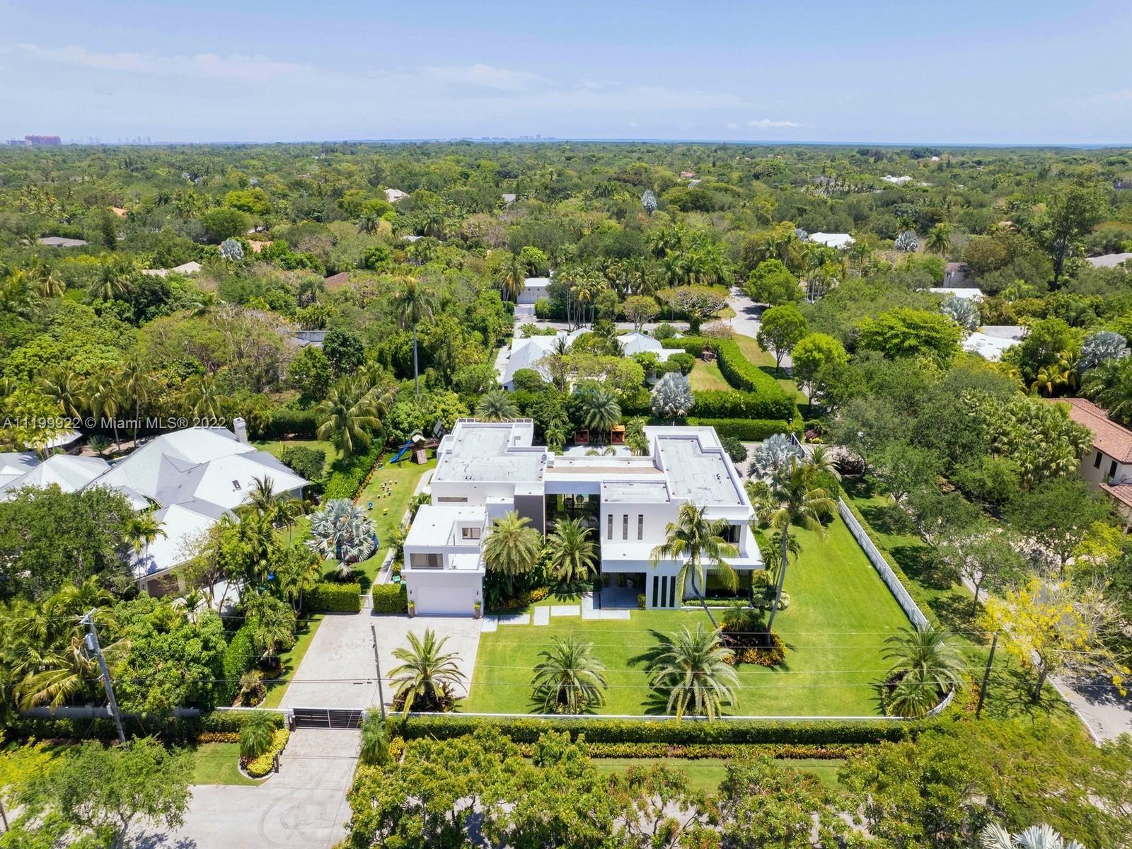 Aerial view of a modern white home with a palm-lined lawn, gated driveway, and dense tropical greenery.