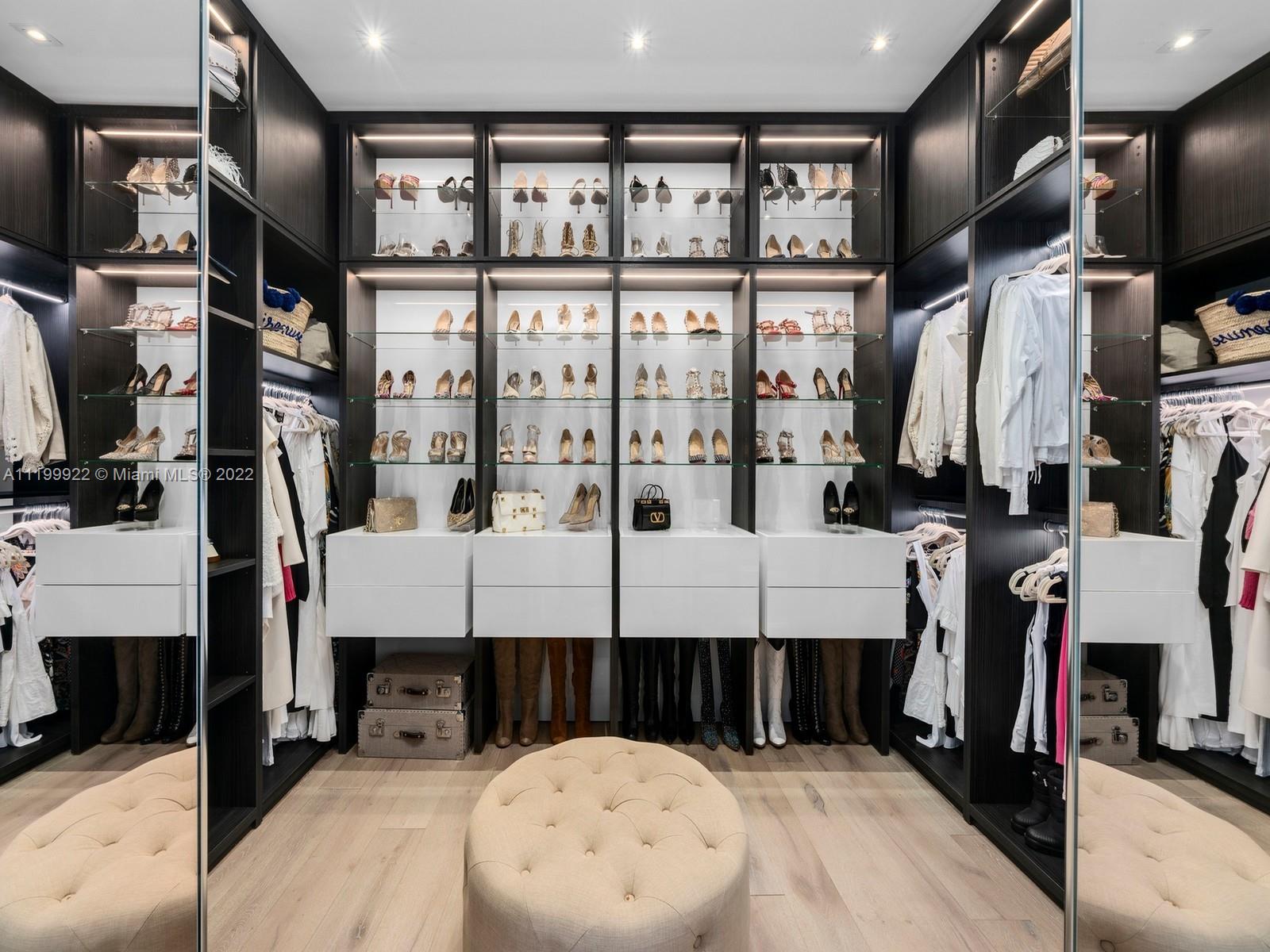 Walk-in closet with illuminated shoe displays, mirrored doors, hanging clothes, and a tufted ottoman.