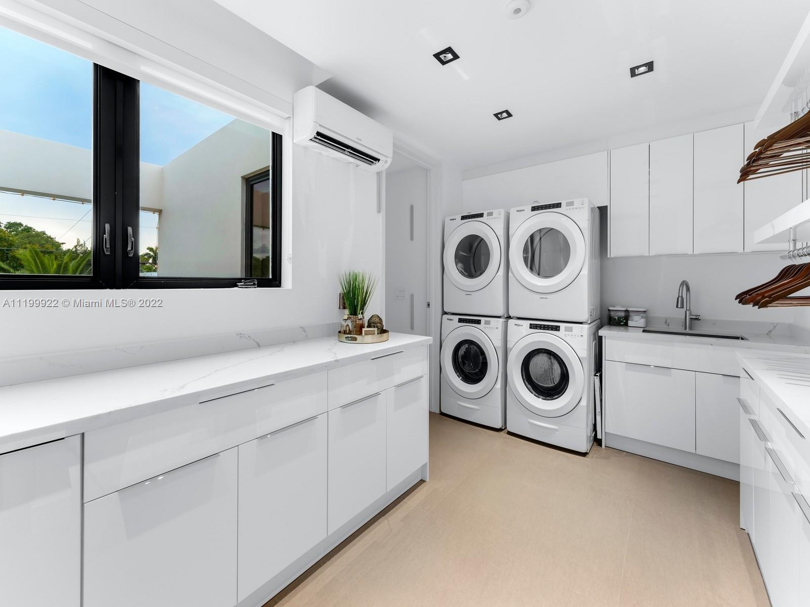 Modern laundry room with stacked front-loading washer and dryer, white cabinetry, and a large window.
