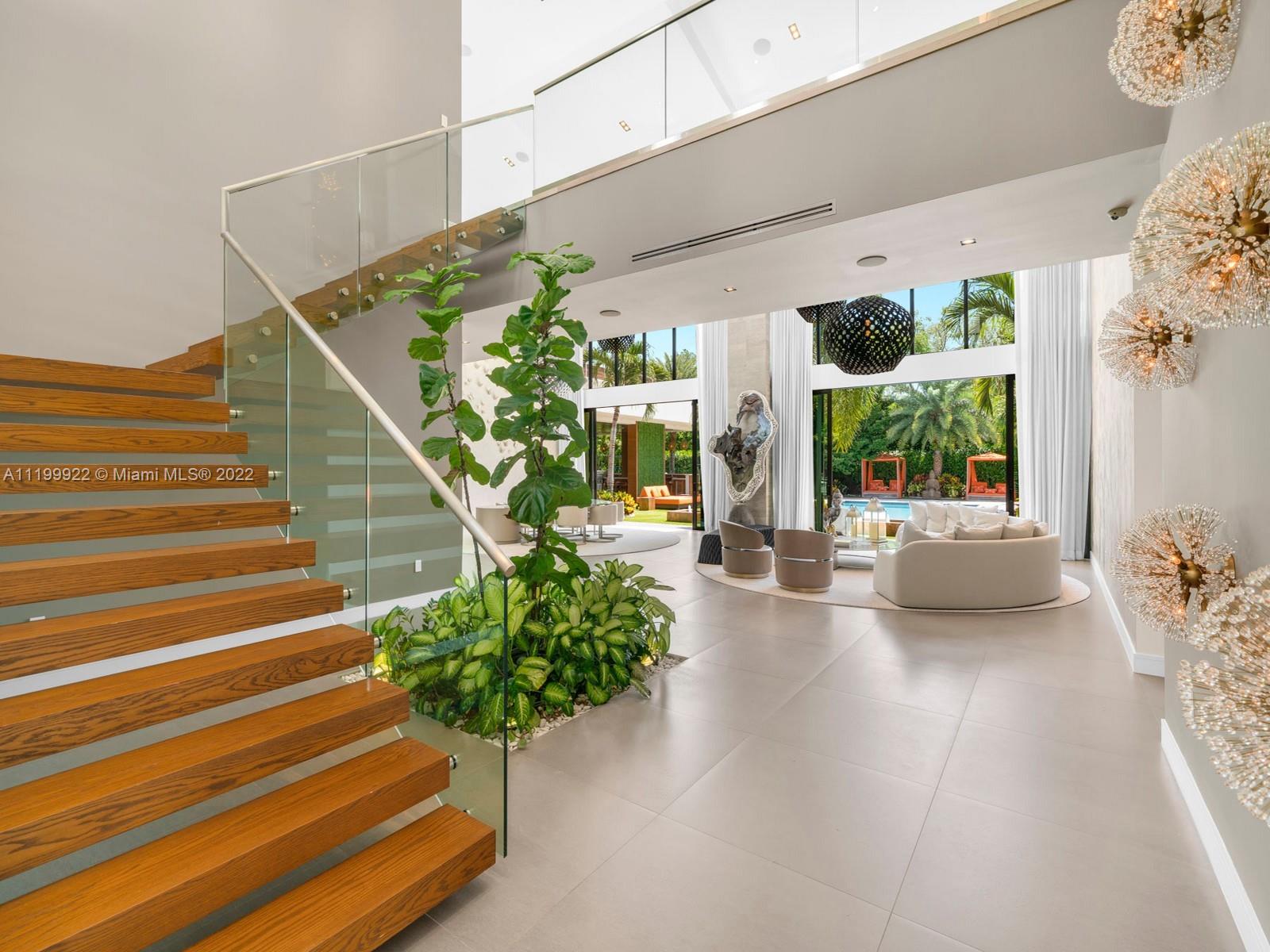 Double-height living room with floating wood stairs, glass railing, and views of a tropical garden and pool.
