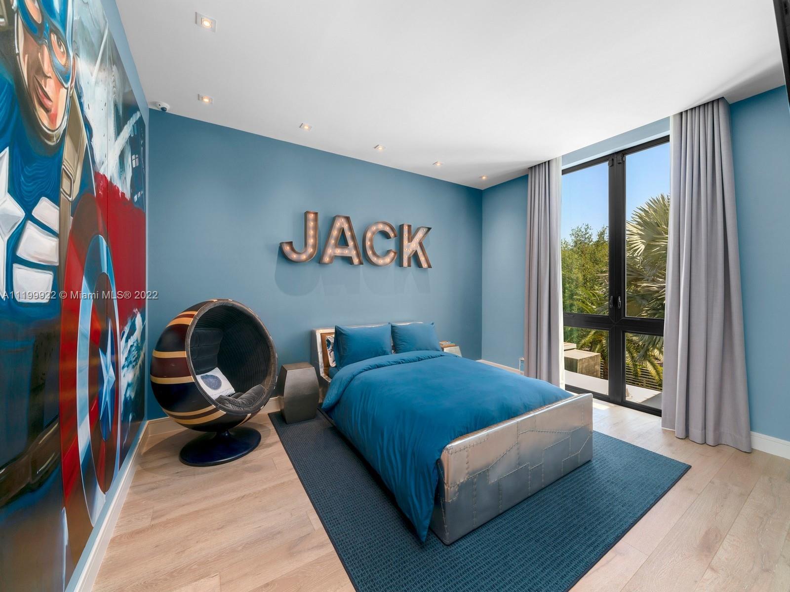 Bedroom with blue bedding, a colorful wall mural, illuminated JACK letters, and tall windows overlooking greenery.
