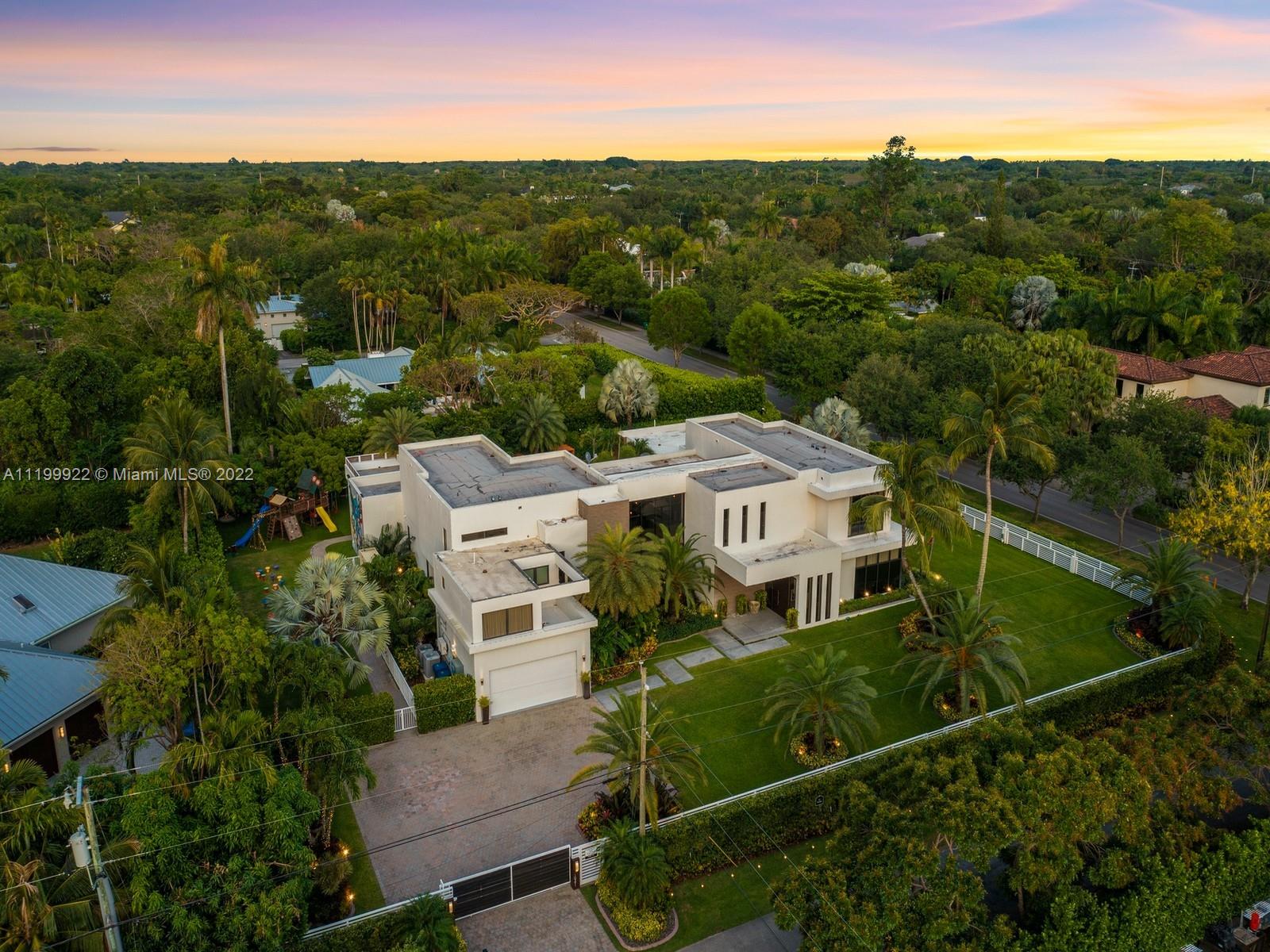 Modern white home with a gated driveway and tropical landscaping, viewed from above at sunset.