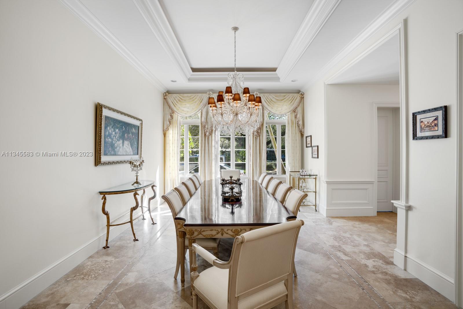 Formal dining room with a long polished table, ornate chandelier, and tall curtained windows.