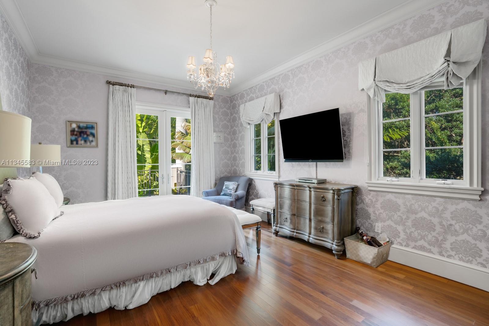 Furnished bedroom with a white bed, chandelier, and windows overlooking lush greenery.