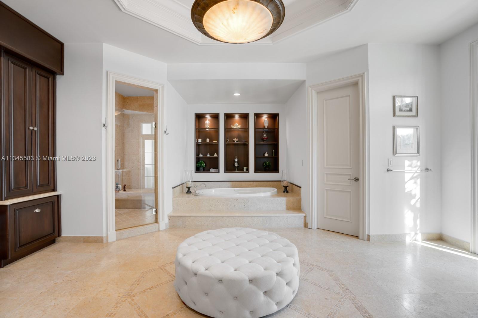 Bedroom with a built-in soaking tub, dark wood cabinetry, and a round tufted ottoman.