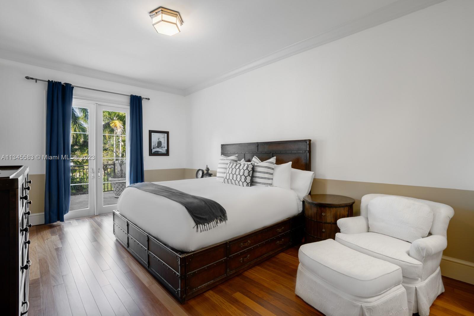 Bedroom with a dark metal bed, blue curtains, balcony doors, and a white armchair with ottoman.