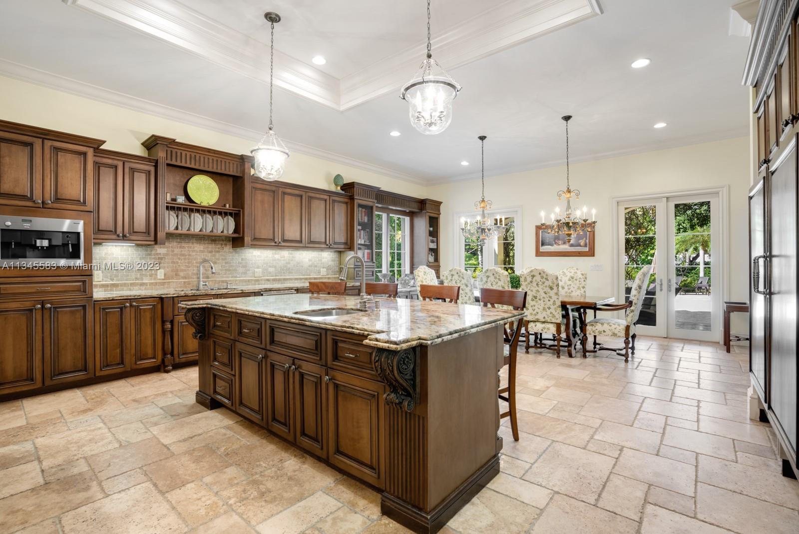 Kitchen with dark wood cabinetry, a large granite island, stone floors, and dining area beside garden-facing French doors.