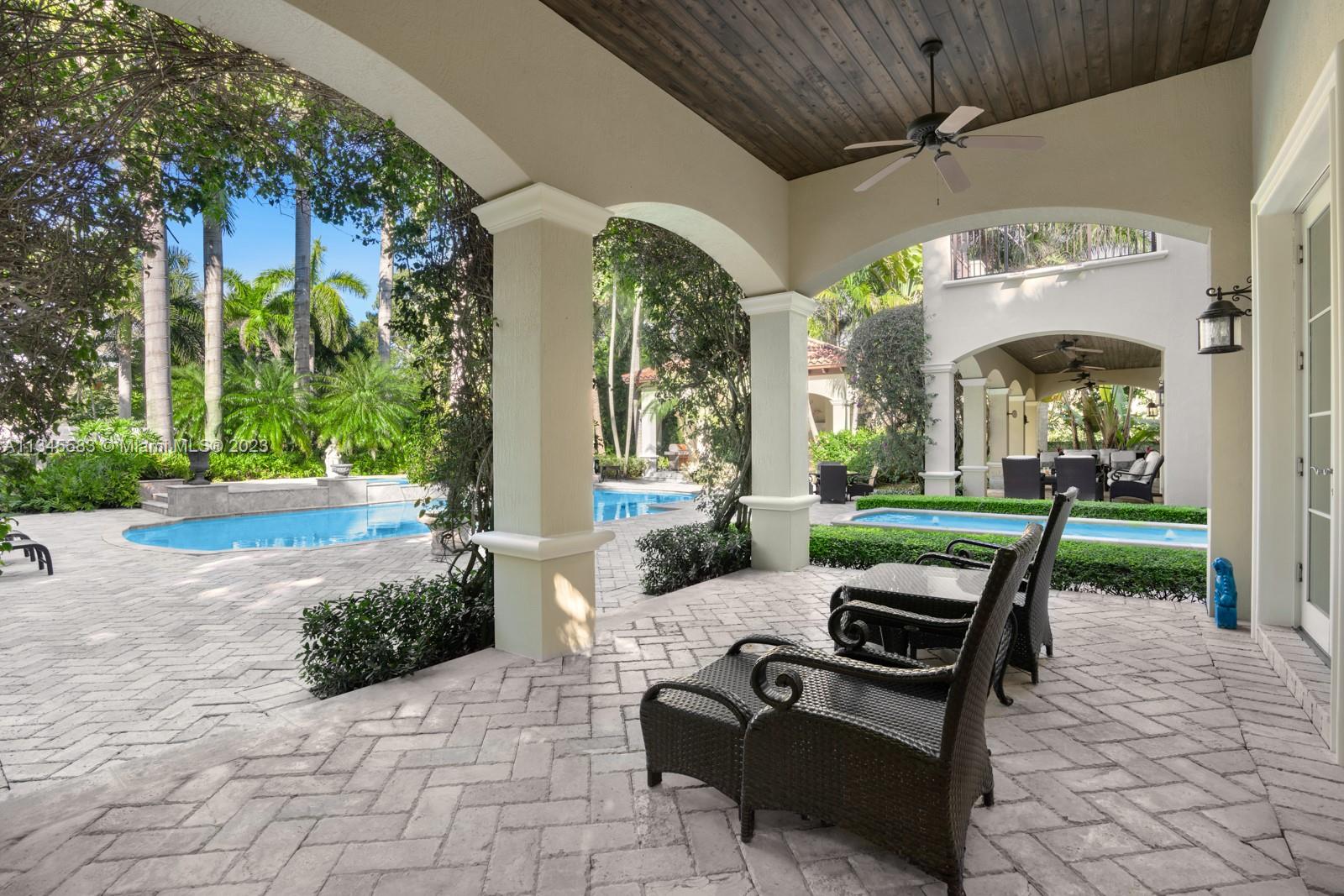 Covered patio with wicker chairs and arched openings overlooking a pool and tropical garden.