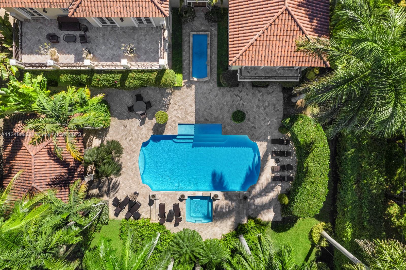 Aerial view of a tropical backyard with a large pool, raised spa, paved patios, and terracotta tile roofs.