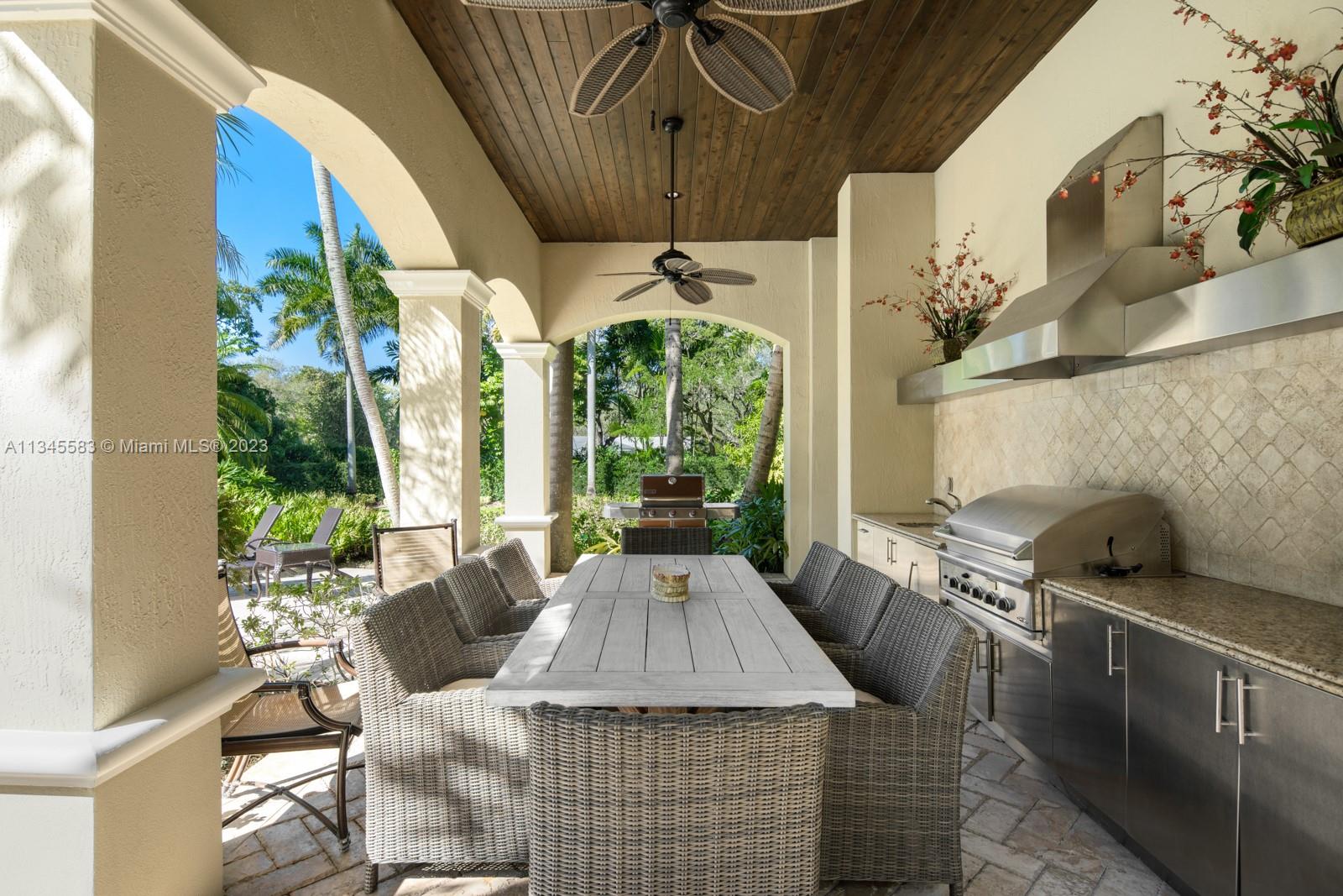 Covered outdoor kitchen and dining patio with wicker seating, a stainless-steel grill, and views of a tropical garden.