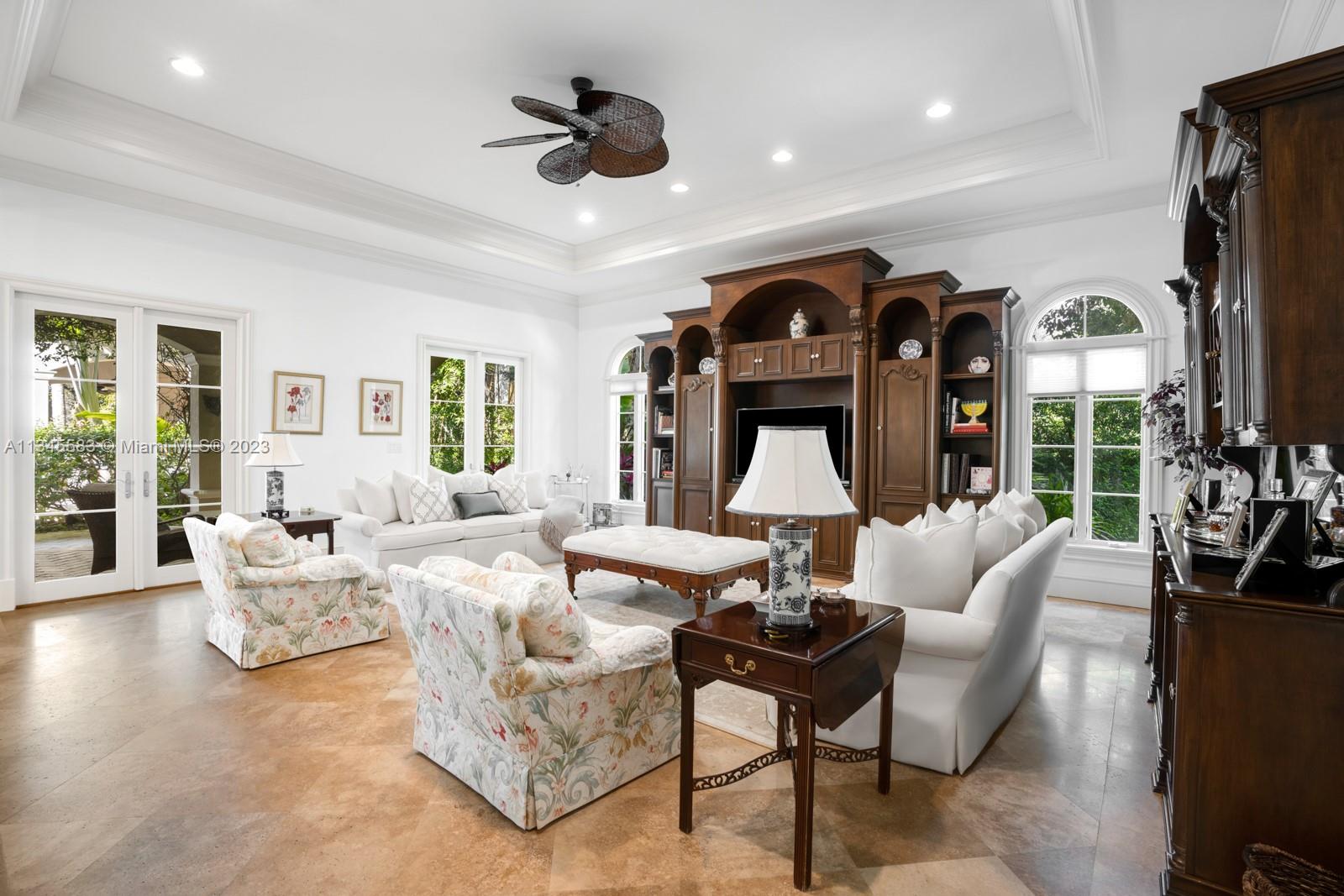 Living room with a dark wood built-in, white seating, floral armchairs, and large windows.