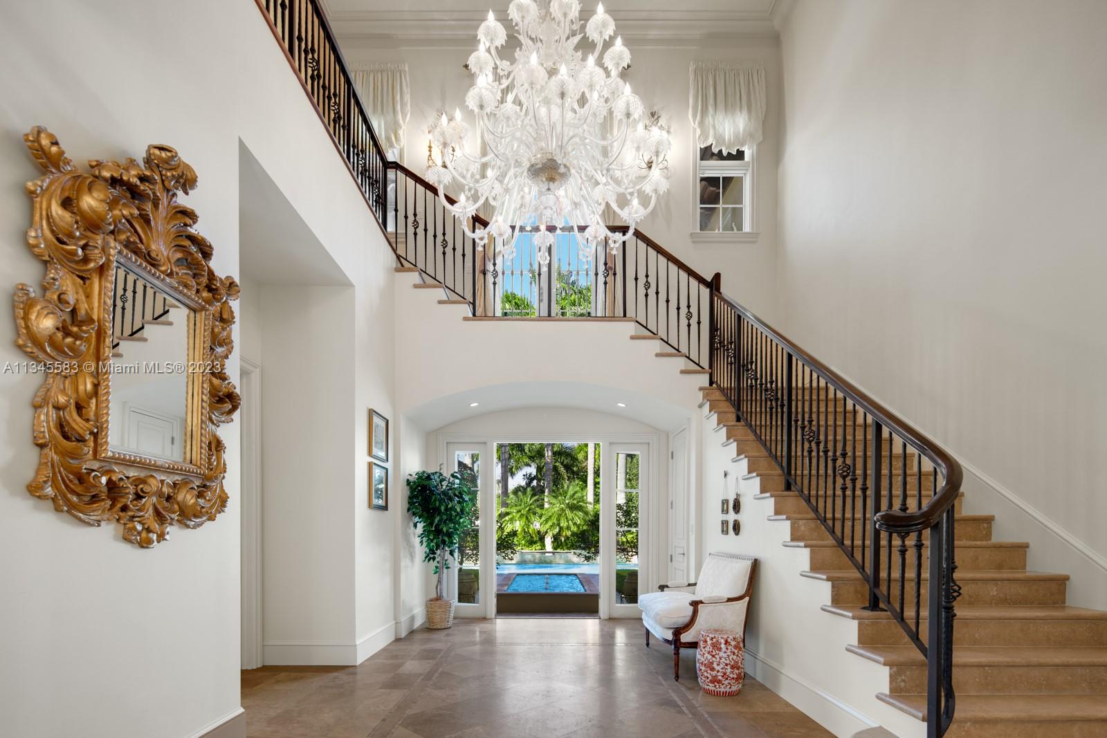 Two-story foyer with a sweeping staircase, oversized chandelier, ornate mirror, and pool visible through glass doors.
