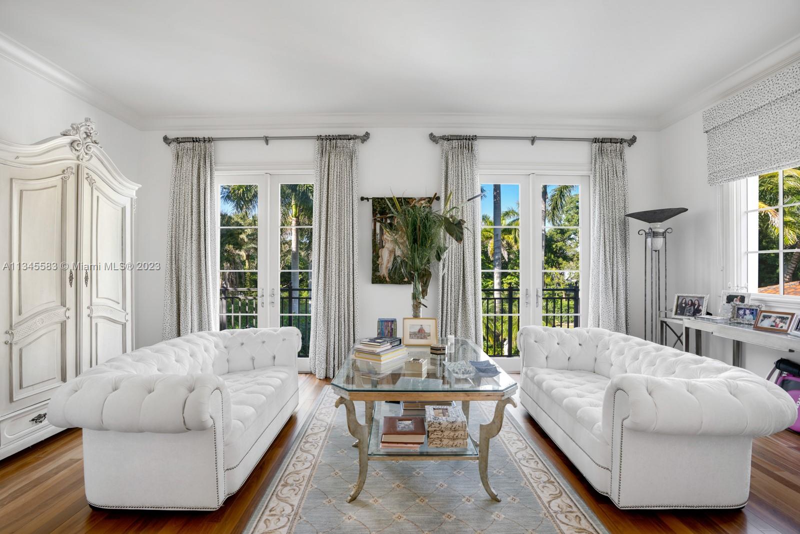 Living room with white tufted sofas, a glass coffee table, and French doors facing palm trees.