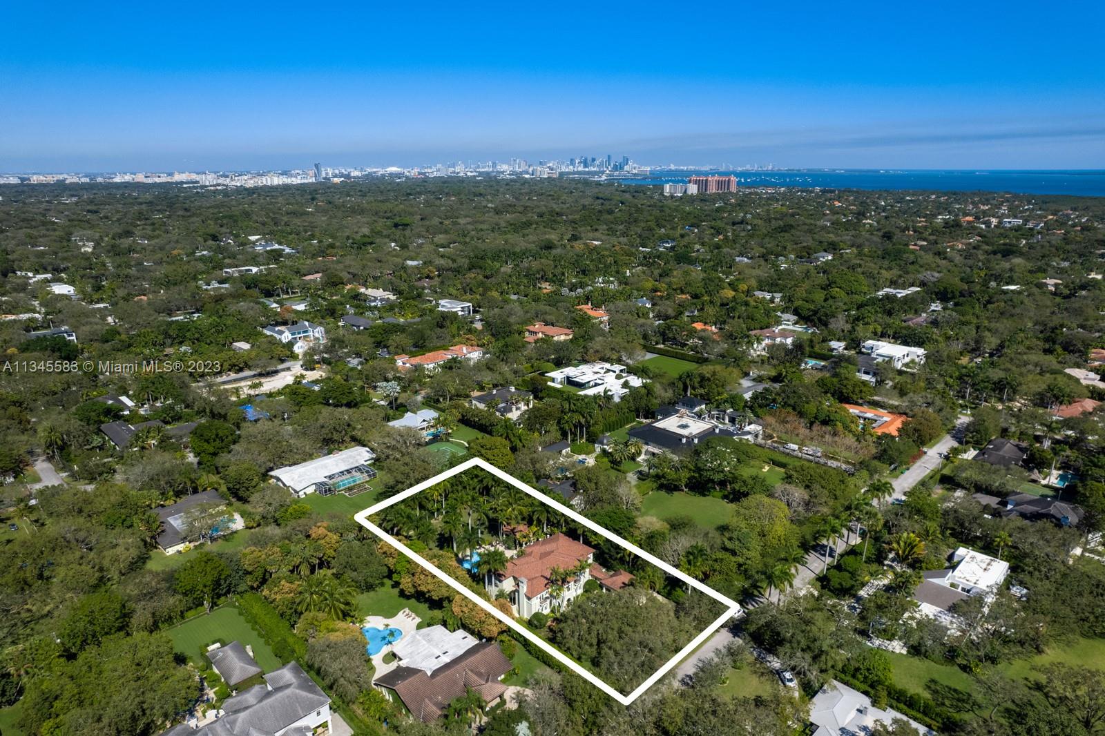 Aerial view of the white-outlined property, surrounded by trees, with the Miami skyline and ocean in the distance.