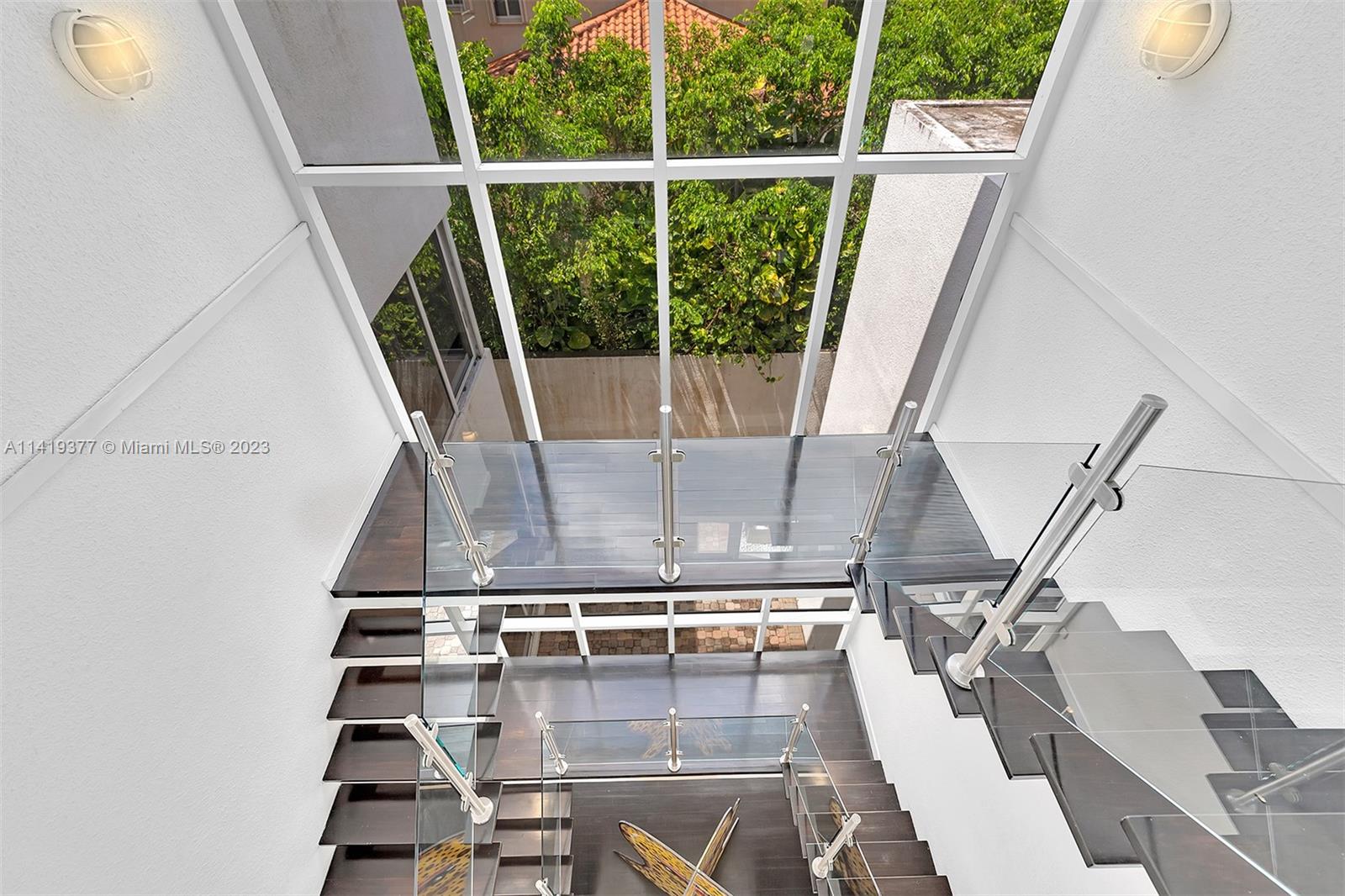 Floating dark staircase with glass railings beneath a tall glass ceiling overlooking greenery.