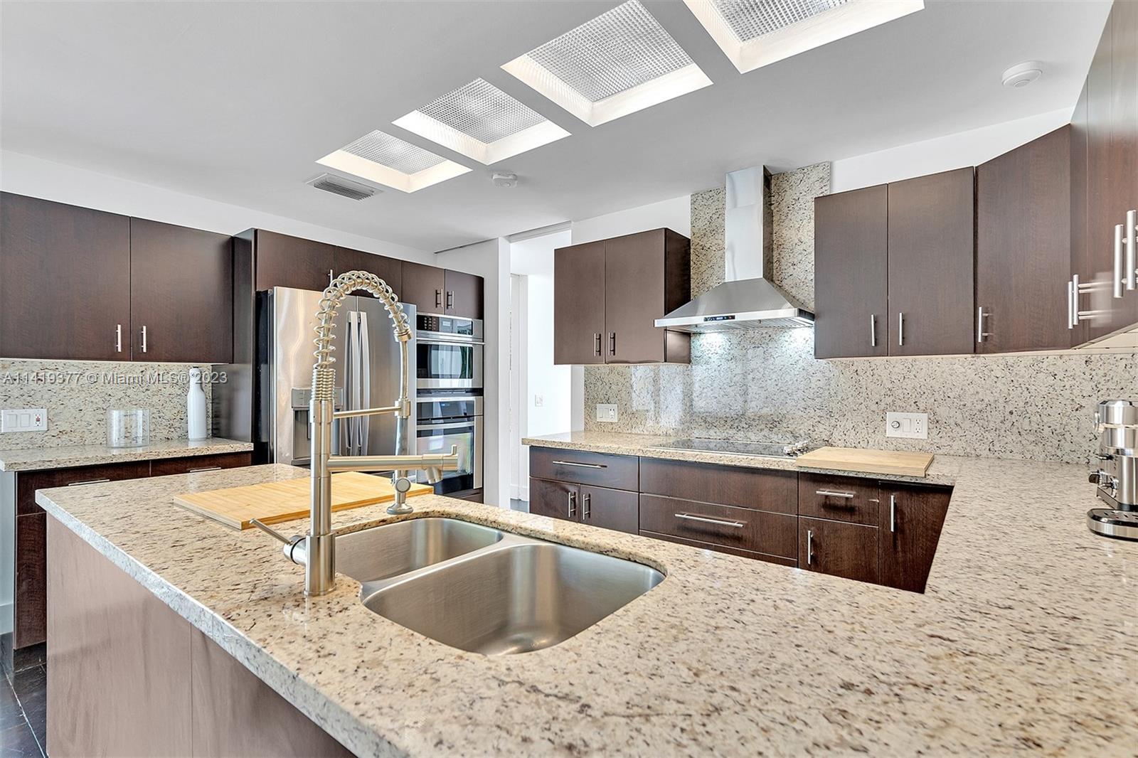 Modern kitchen with dark wood cabinets, granite counters, a large double-sink island, and stainless steel appliances.