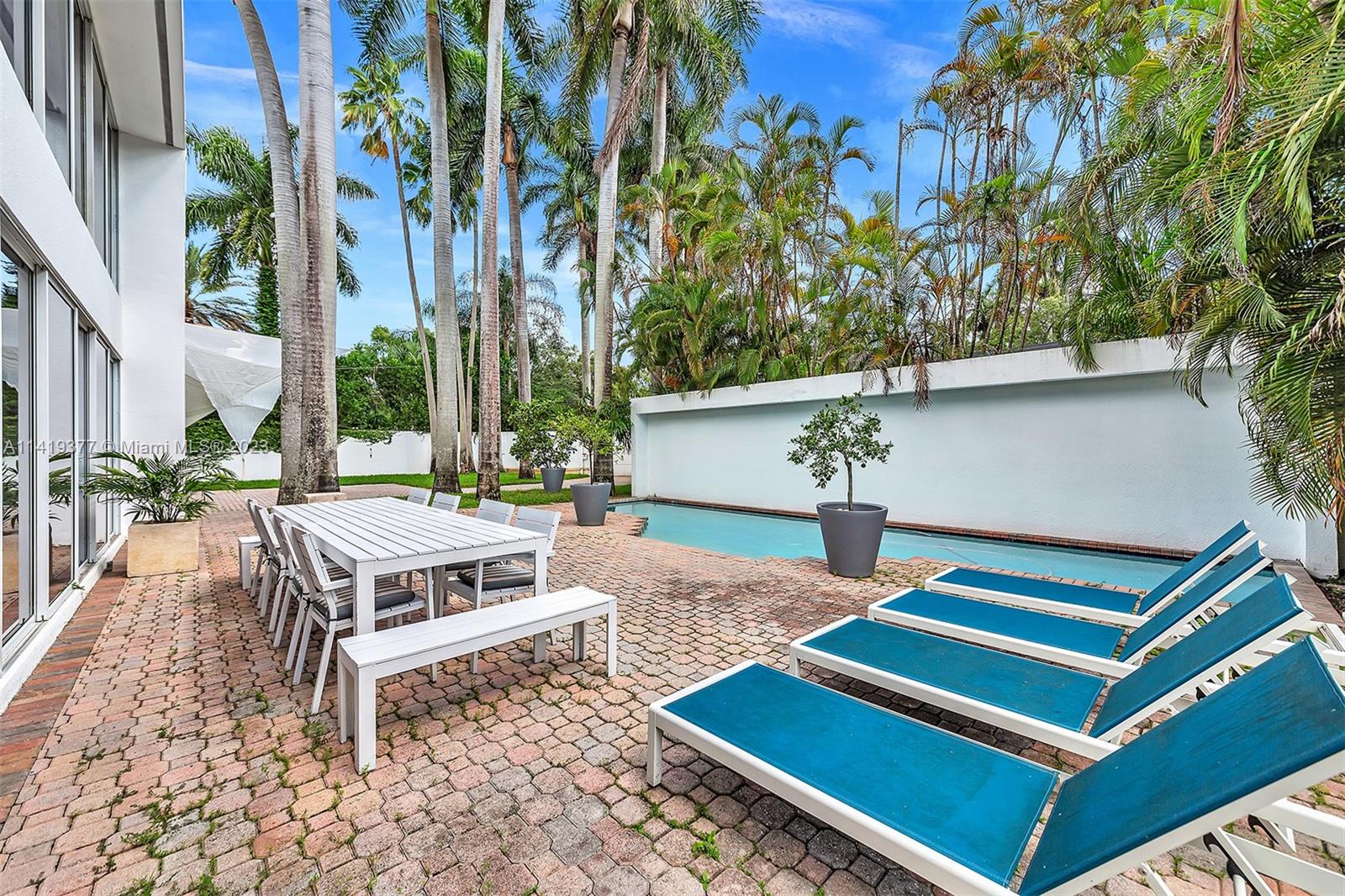 Narrow backyard pool beside a brick patio with blue loungers, white dining furniture, and tall palms.