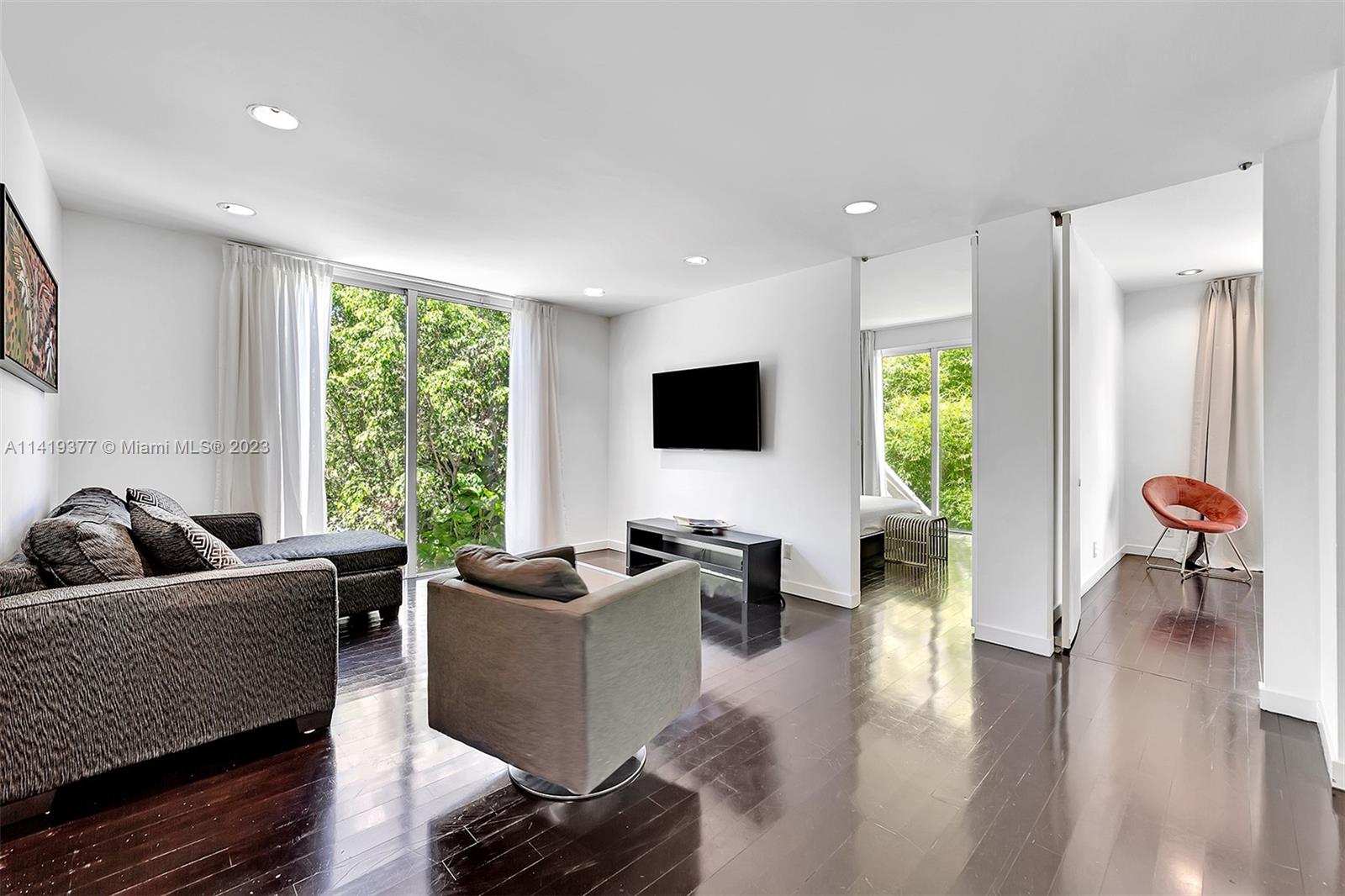 Living room with gray seating, glossy dark floors, and large windows overlooking greenery.