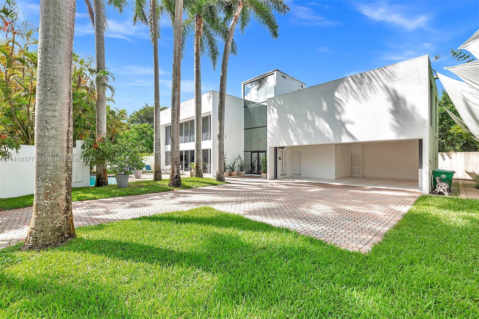 Modern white home with a glass entry, open carport, palm-lined paver driveway, and lawn.