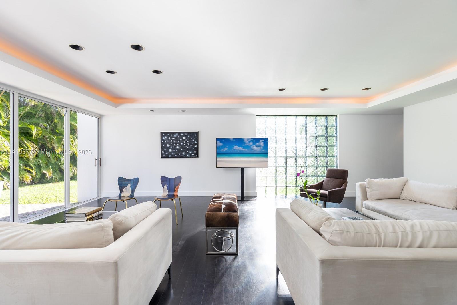 Bright living room with white sofas, warm ceiling lighting, and sliding glass doors facing a leafy garden.