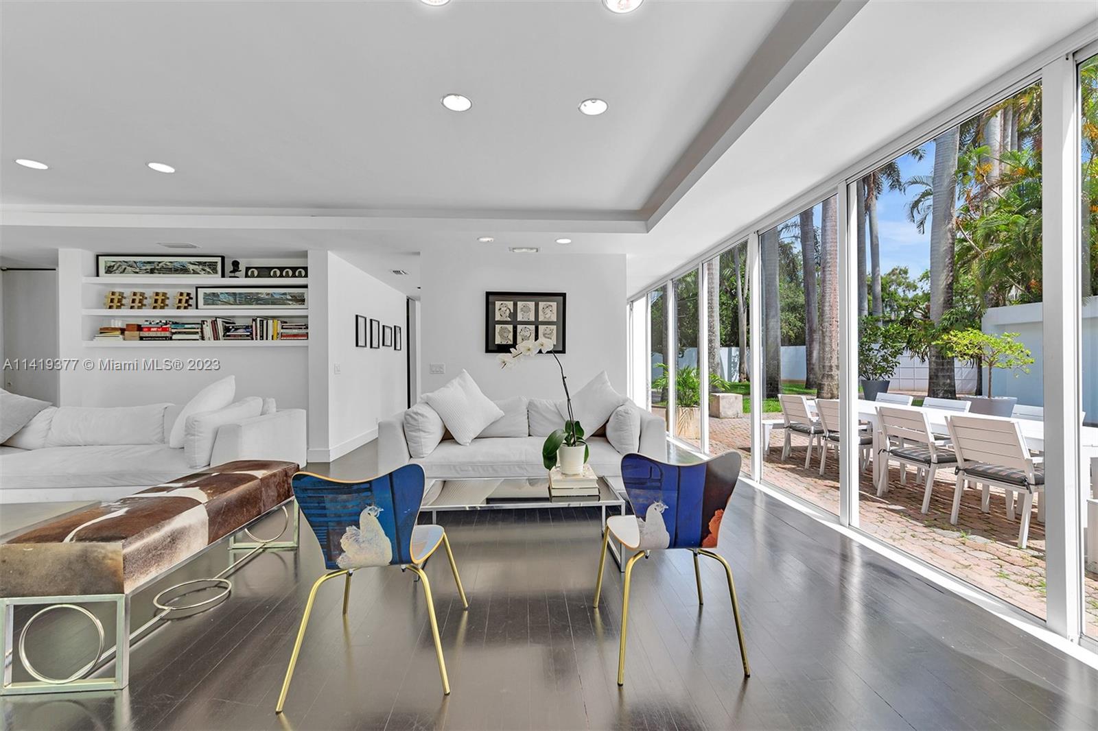 Living room with white sofas and blue chairs beside glass doors opening onto a palm-lined patio.