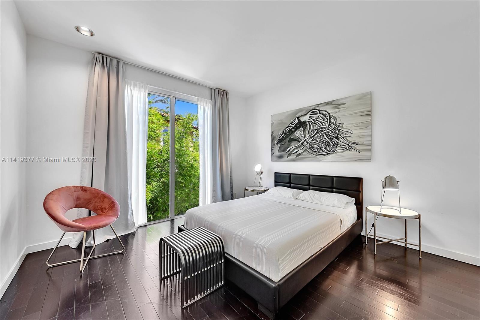 Furnished bedroom with a black platform bed, pink accent chair, and sliding glass doors overlooking greenery.