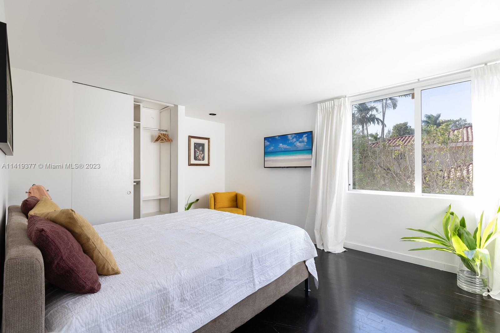 Bright bedroom with a white bed, open closet, yellow chair, and large window overlooking greenery.