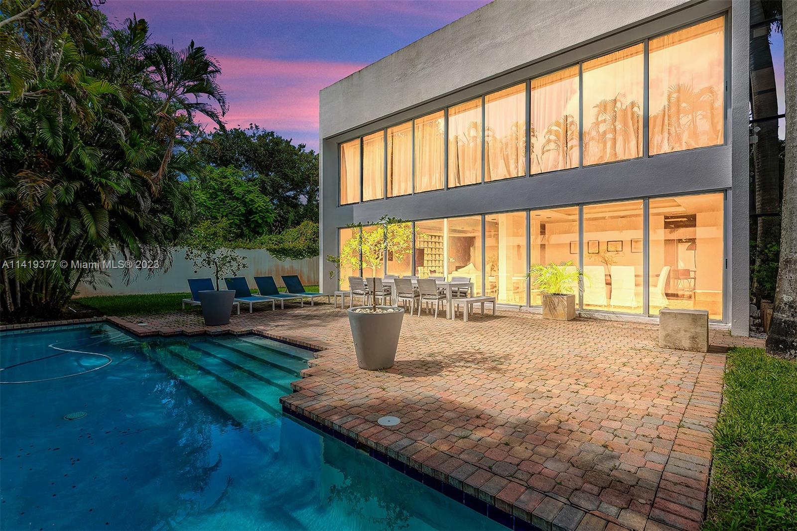 Modern glass-fronted home beside a swimming pool and brick patio at dusk.