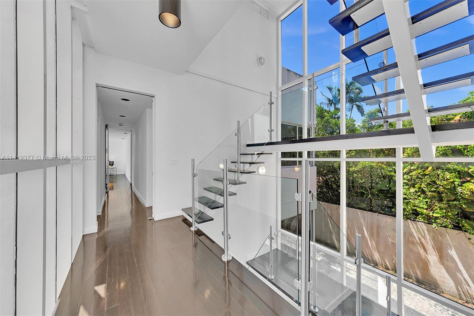 Modern hallway with a glass-railed floating staircase and tall windows overlooking greenery.