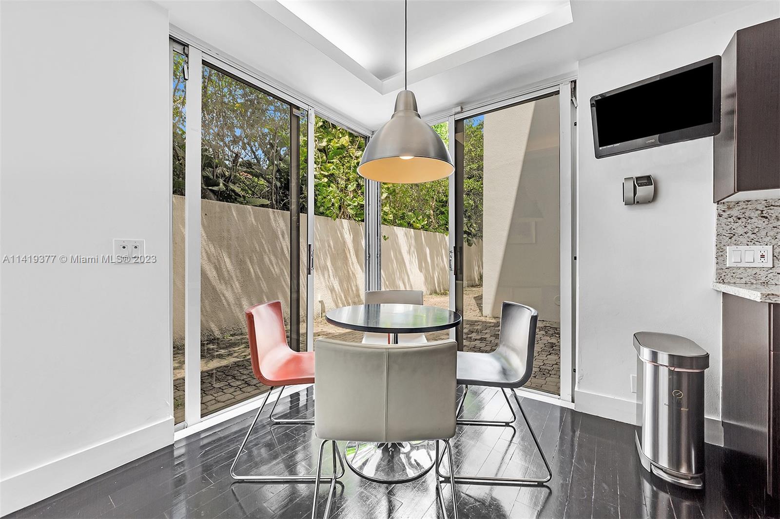Dining area with a round table and chairs beside glass doors opening to a leafy courtyard.