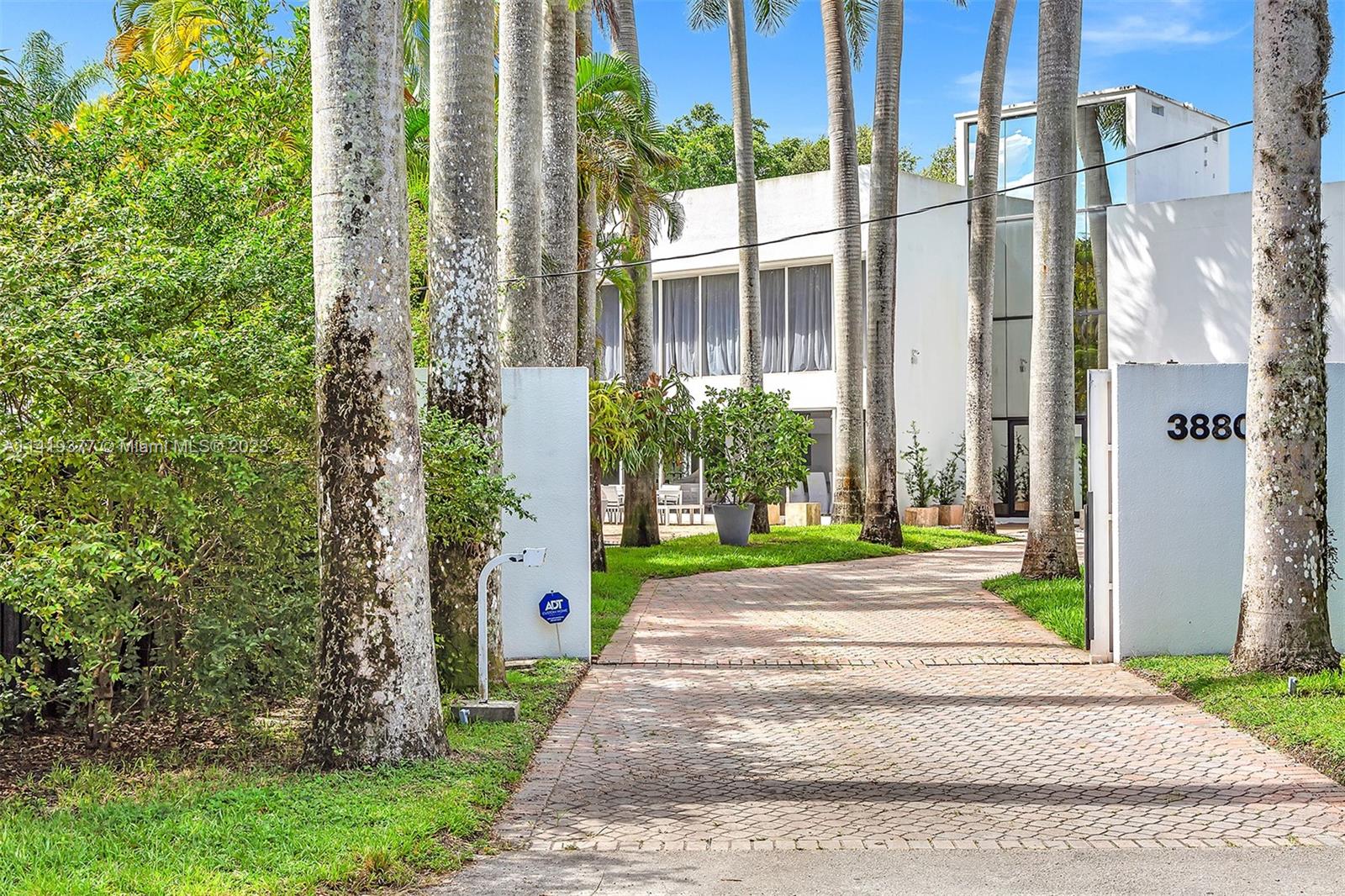 Modern white home behind tall palms, with a brick driveway and gated entrance marked 3880.