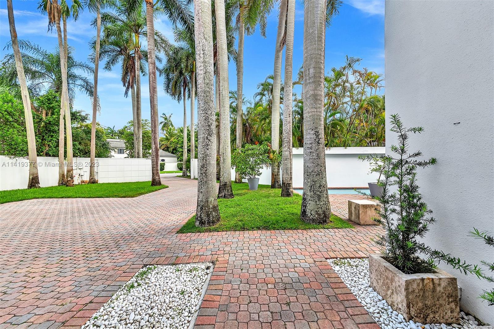 Brick-paved courtyard lined with tall palms, with tropical landscaping and a glimpse of the pool.