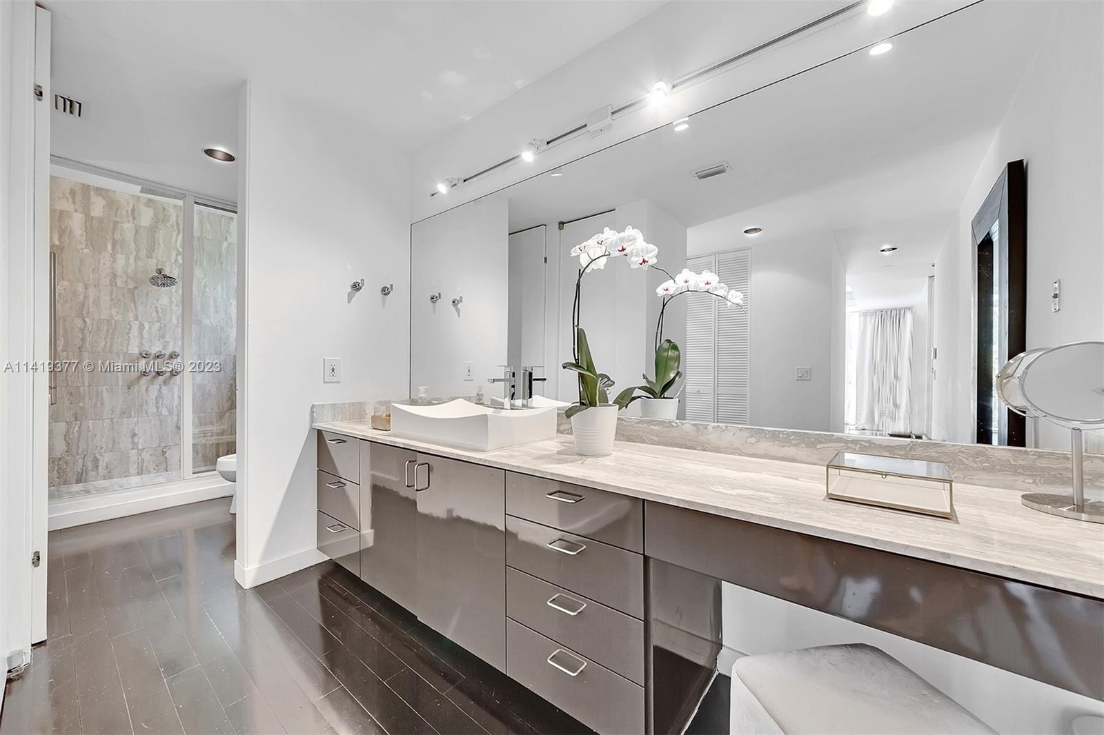 Modern bathroom with a long gray vanity and vessel sink beside a glass-enclosed stone shower.