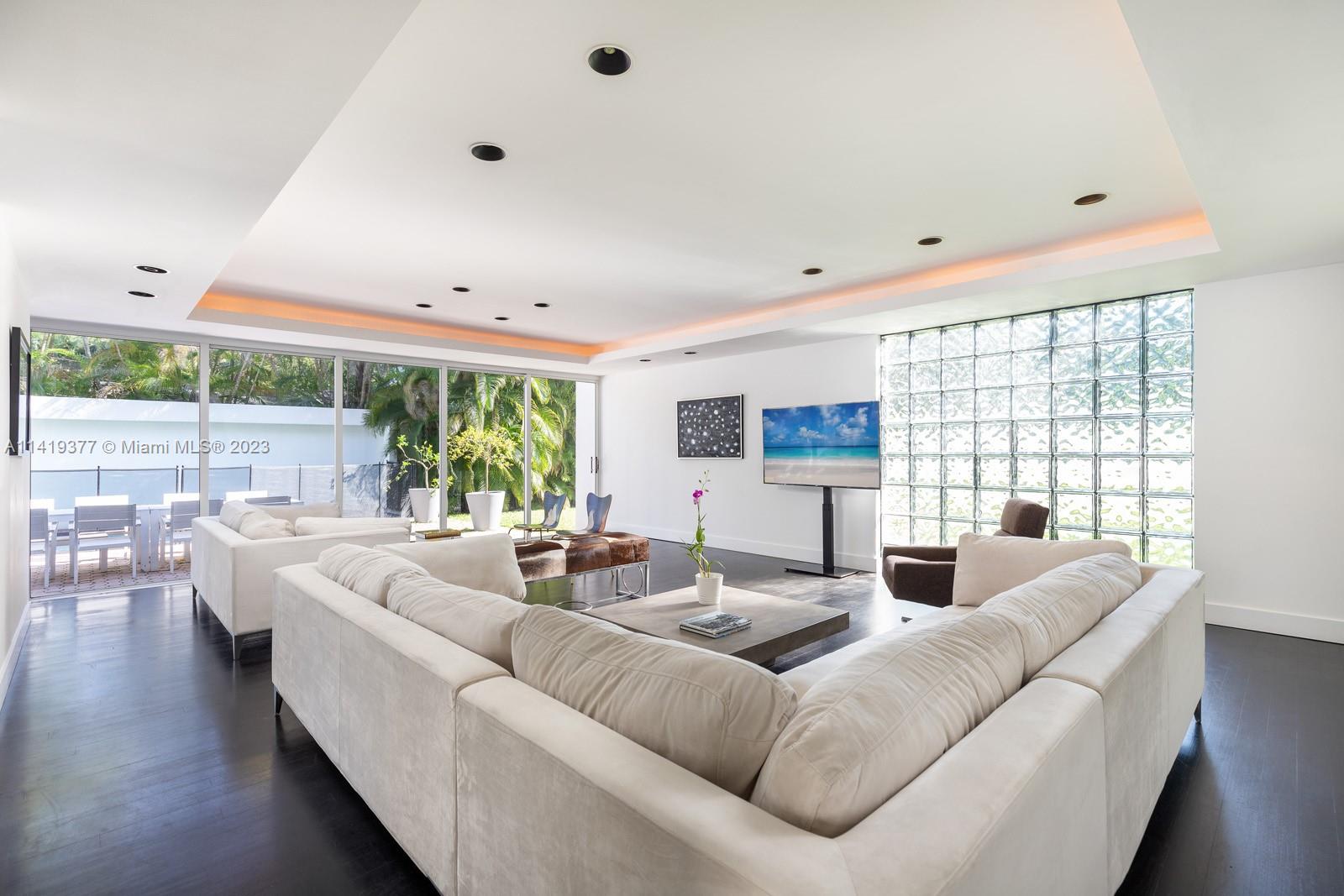 Modern living room with white sectional sofas, glass-block wall, and doors opening to a poolside patio.