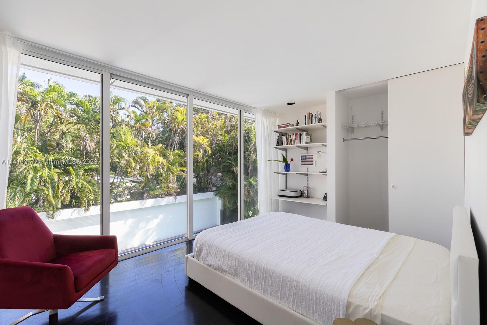 Bedroom with floor-to-ceiling glass doors overlooking tropical greenery, a white bed, and burgundy chair.