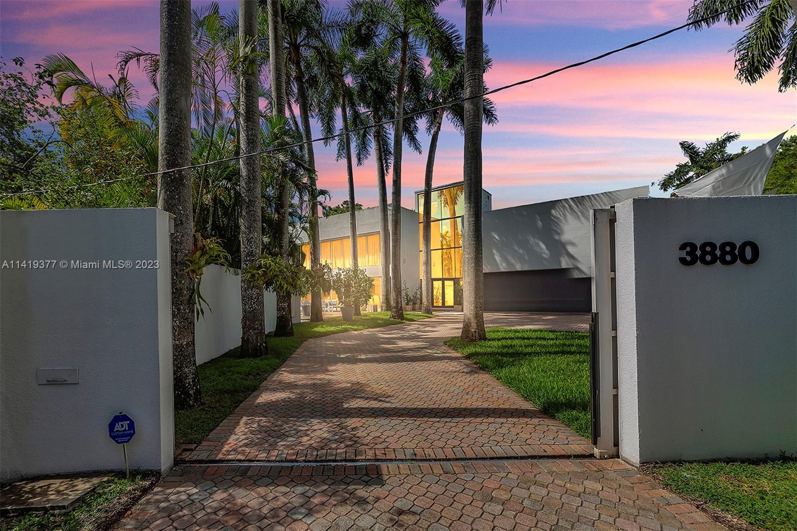 Modern white home behind a palm-lined brick driveway and gate marked 3880 at dusk.