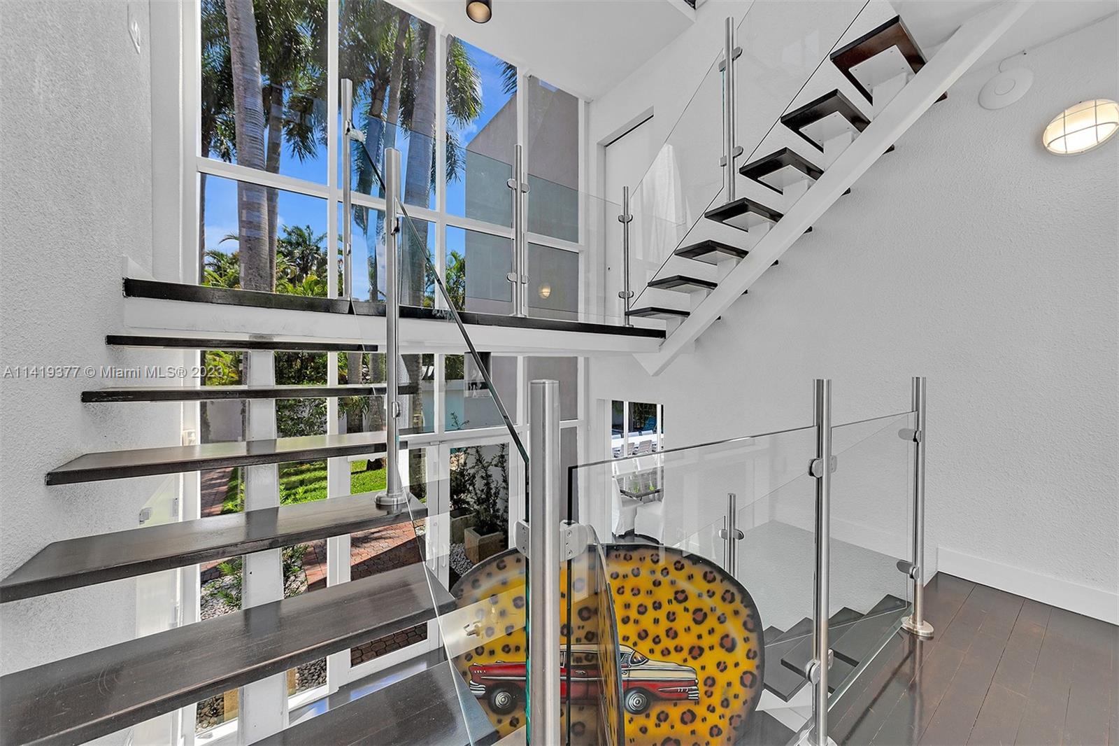Double-height stairwell with floating dark wood stairs, glass railings, and a colorful circular car artwork.