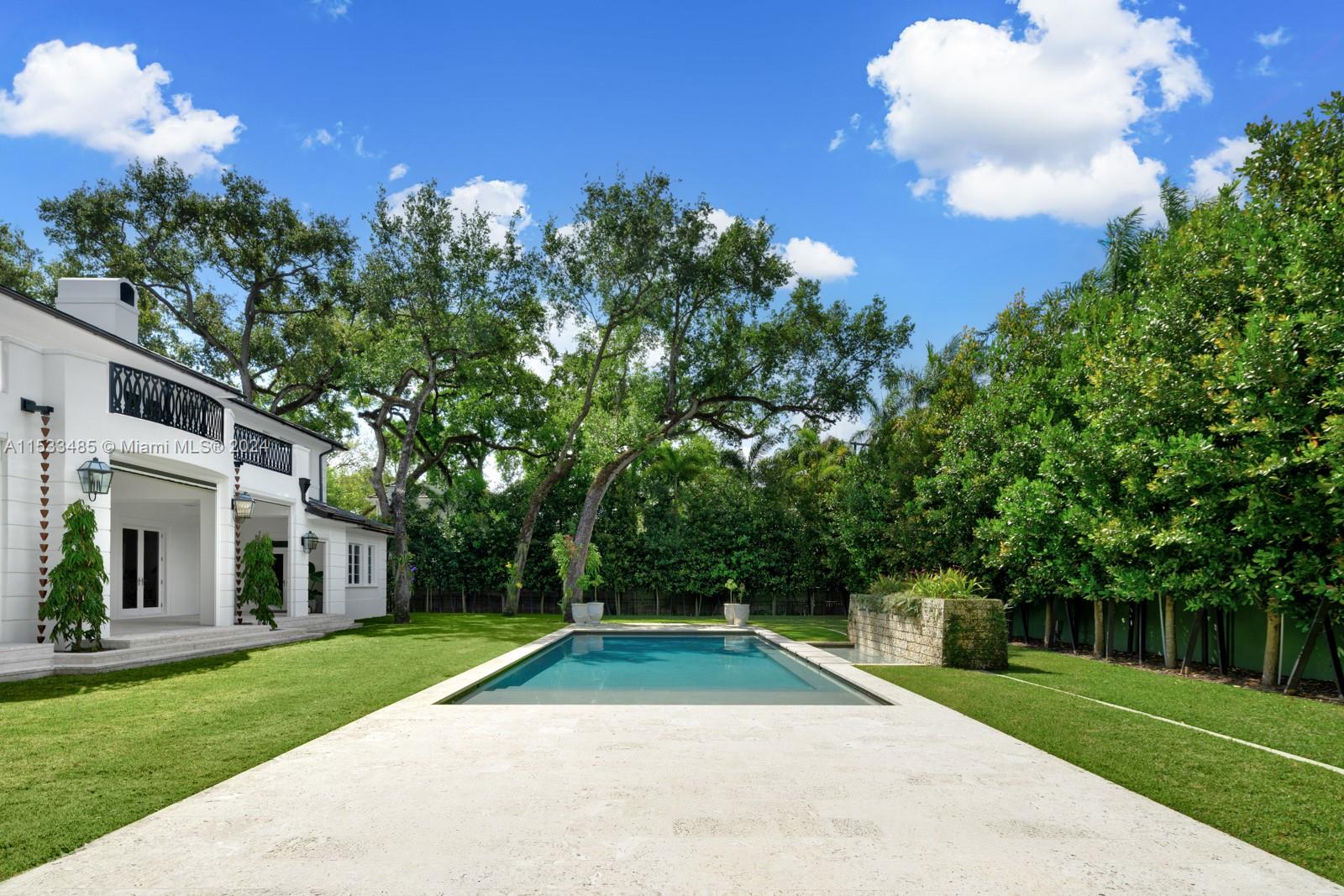 Backyard swimming pool beside a white two-story home, framed by a lawn, mature trees, and privacy hedges.