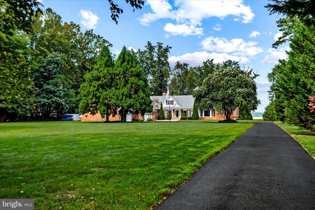 White and brick house beyond a broad lawn, framed by mature trees, with a long driveway and water in the distance.